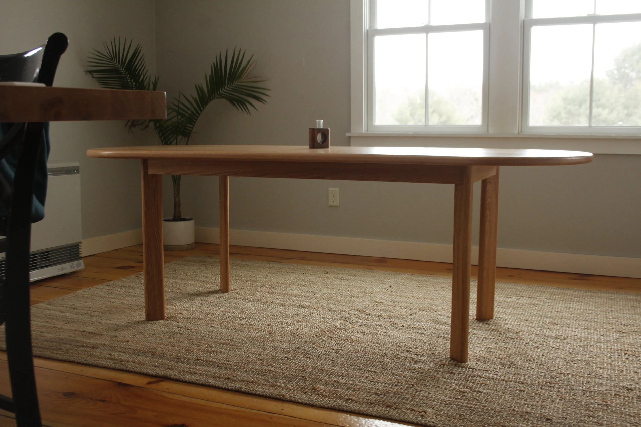 A wooden dining table with rounded edges in a room with large windows, a potted palm tree, and a beige woven rug on a wooden floor.