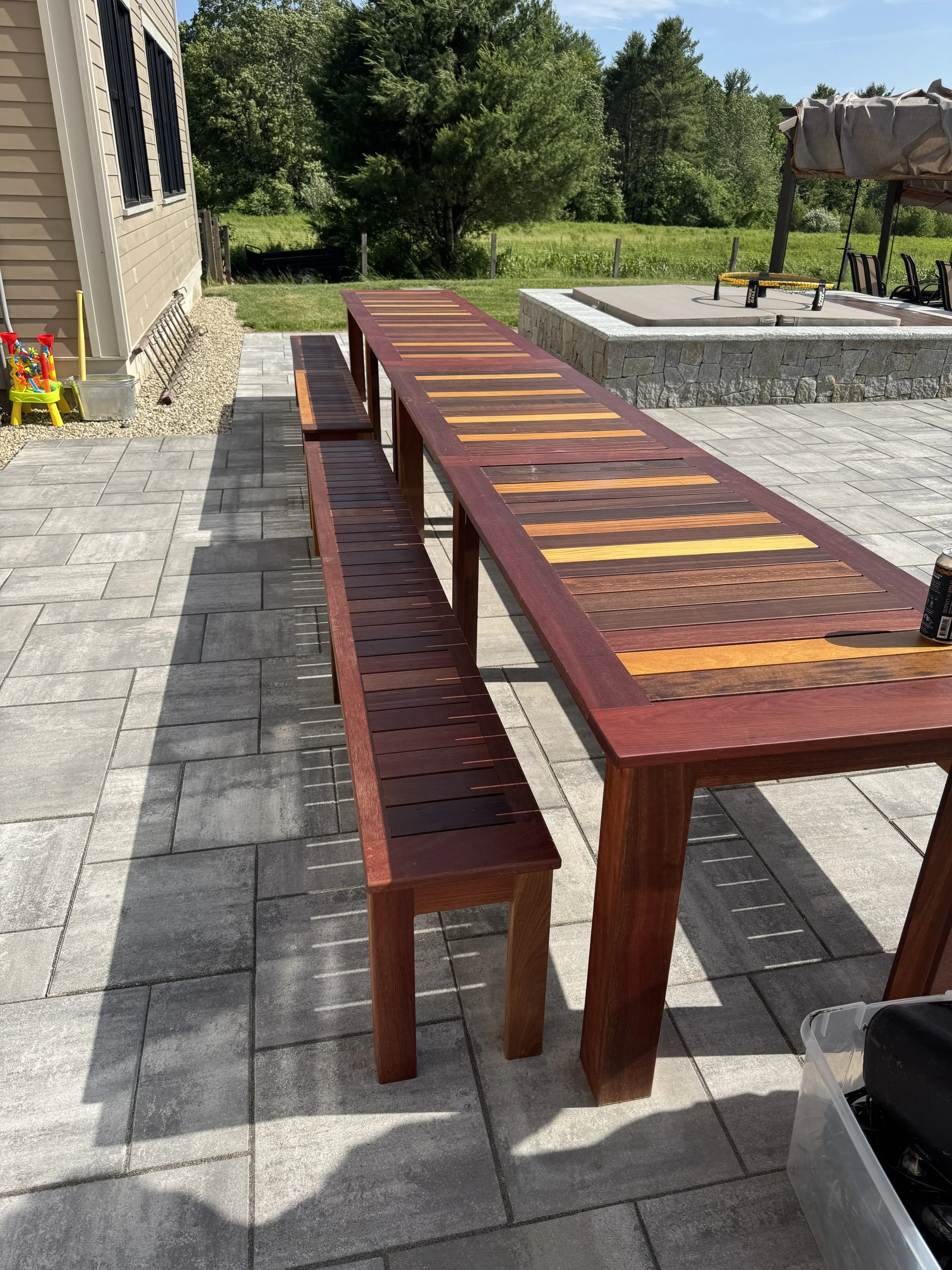 A wooden outdoor table and benches on a stone patio in a backyard. In the background, there is a fire pit area with chairs and a canopy, surrounded by greenery and trees.