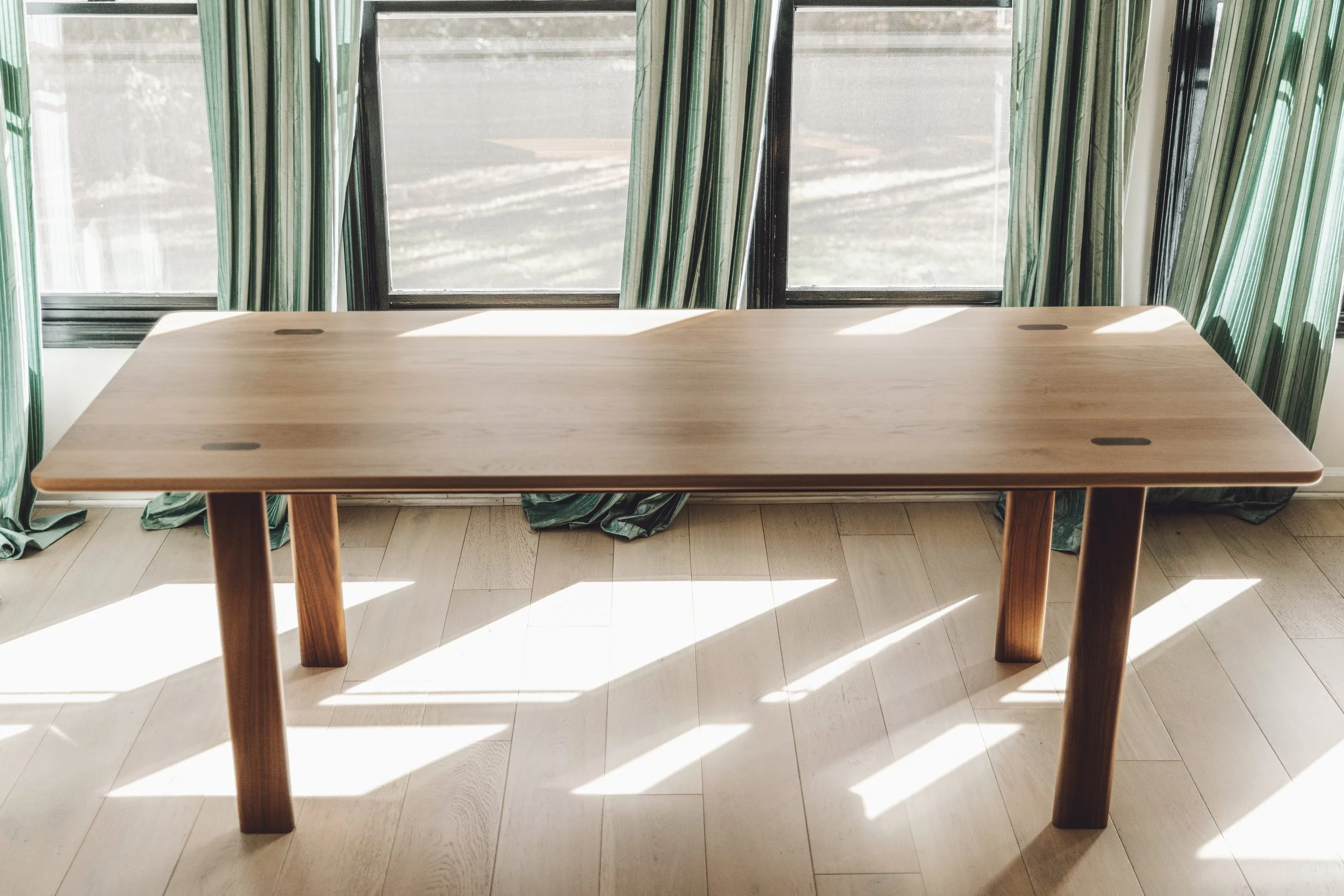 Empty wooden table near large windows with green curtains, sunlight casting shadows on the floor.
