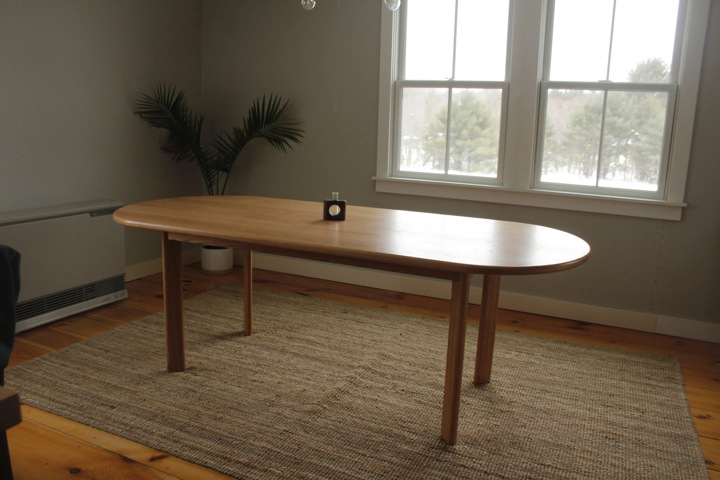 Empty wooden dining table with a small black candle holder on top, placed on a beige rug in front of a large window with a view of trees outside, in a room with beige walls and a potted plant in the corner.