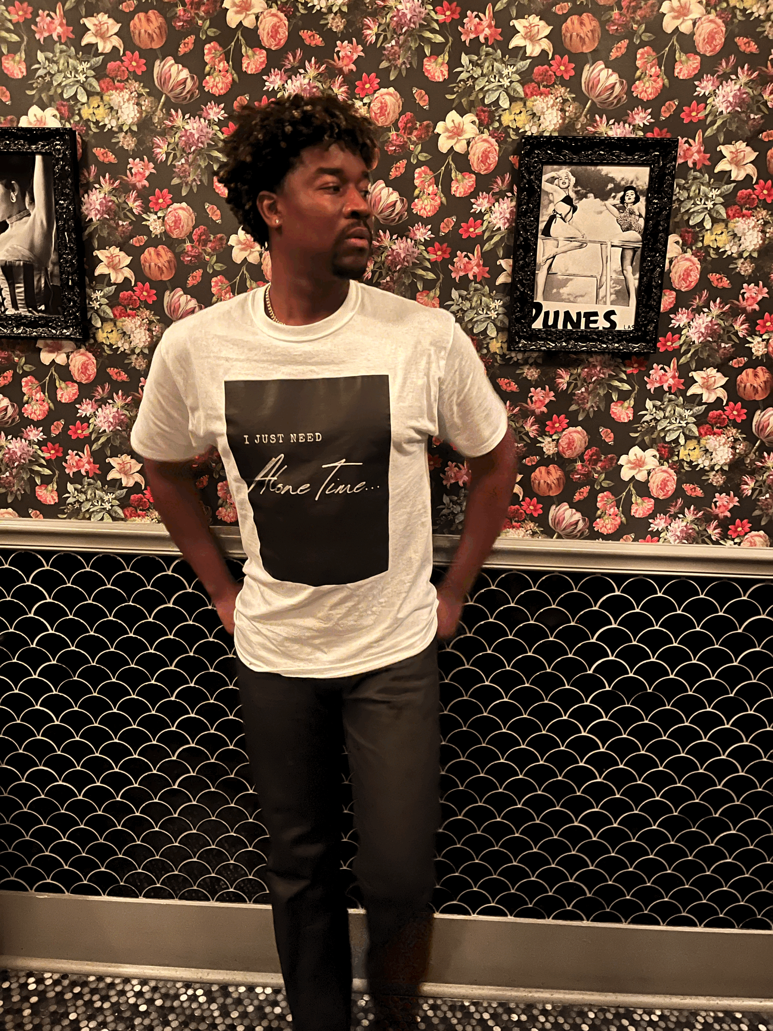 Joseph Valenté posing in the Black and White Alone Time T shirt against a floral wall in The Whale Chicago. The shirt has a graphic that says I Just Need Alone Time