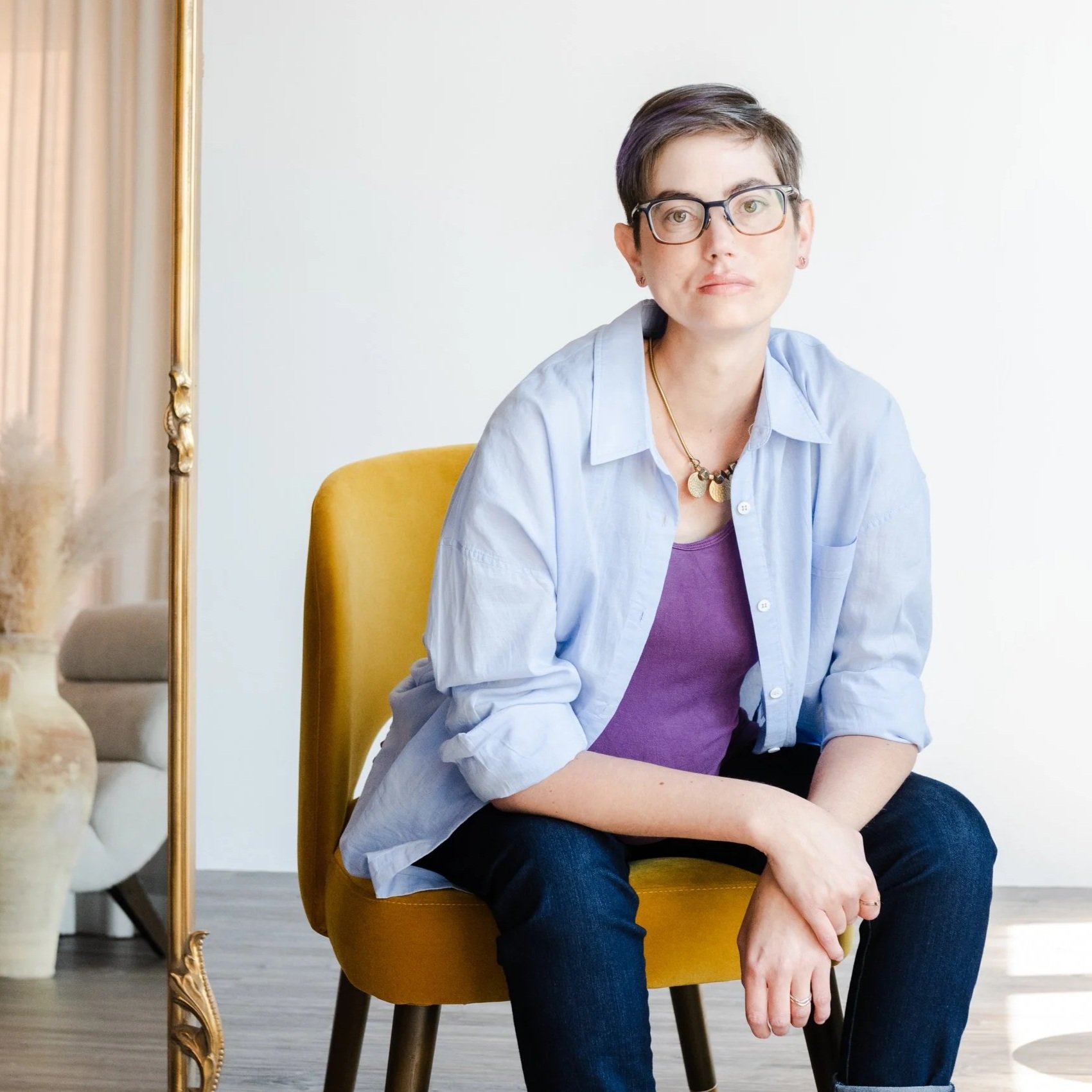 Michelle, a caucasian woman with short dark hair and glasses leaning forward with her elbows on her knees and hands crossed. She is wearing jeans, an open button down shirt and a purple tank top while sitting on a dark yellow fabric chair.