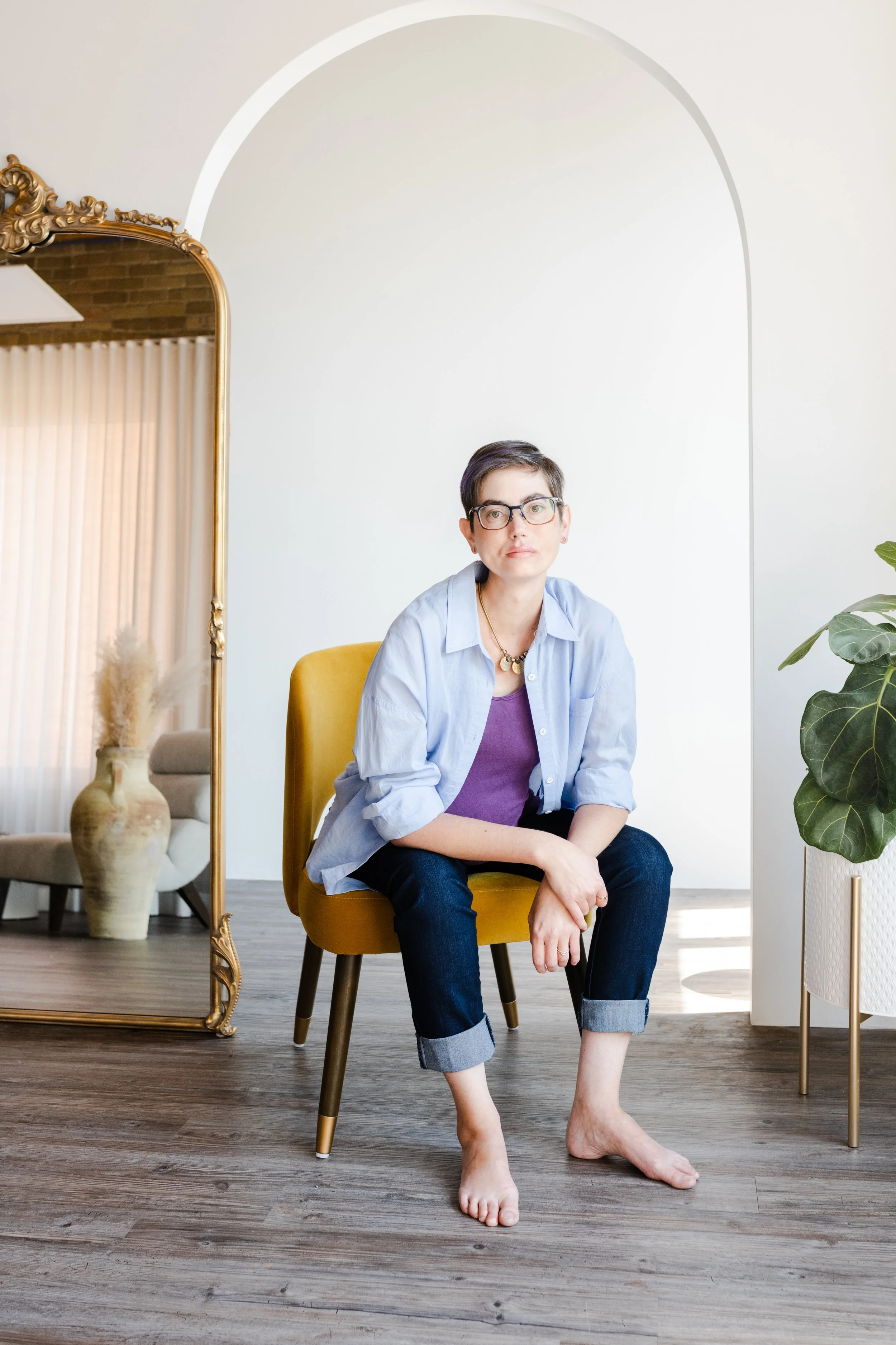 Michelle, a caucasian woman with short dark hair and glasses leaning forward with her elbows on her knees and hands crossed. She is wearing jeans, an open button down shirt and a purple tank top while sitting on a dark yellow fabric chair.