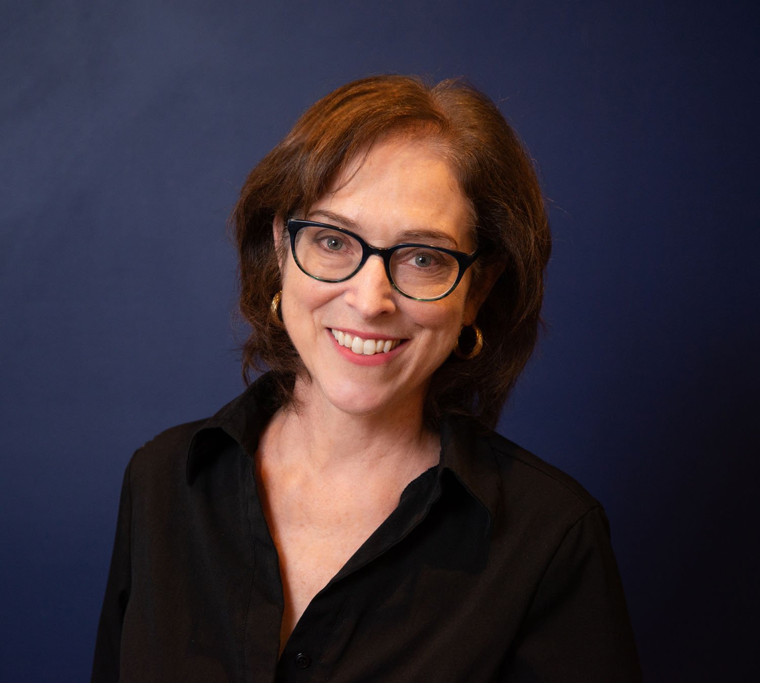 A woman with brown hair, glasses, and gold earrings smiling in front of a dark blue background.