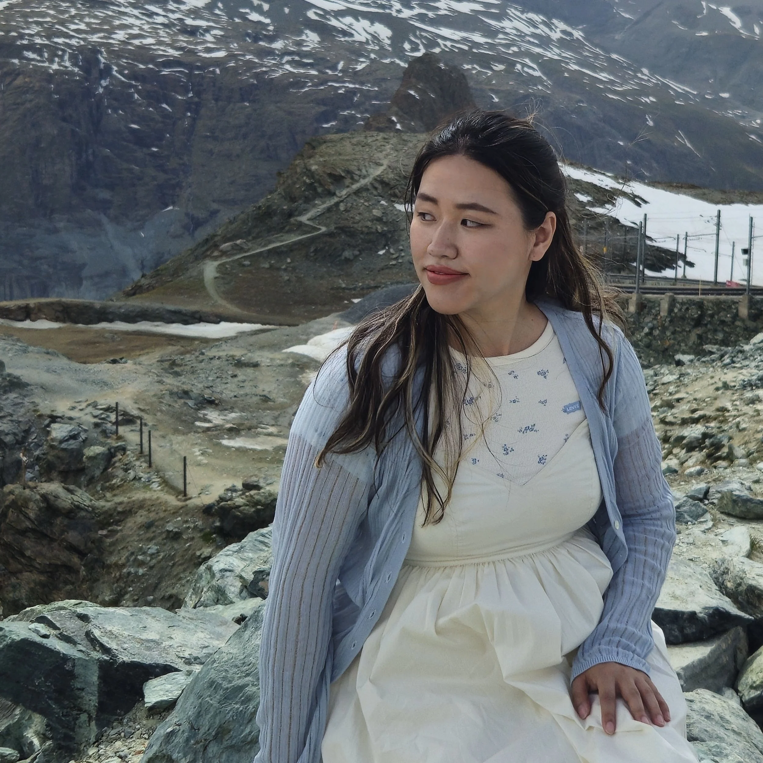 A woman sitting on rocks in a mountainous landscape with snow patches, rocky terrain, and a mountain trail in the background.