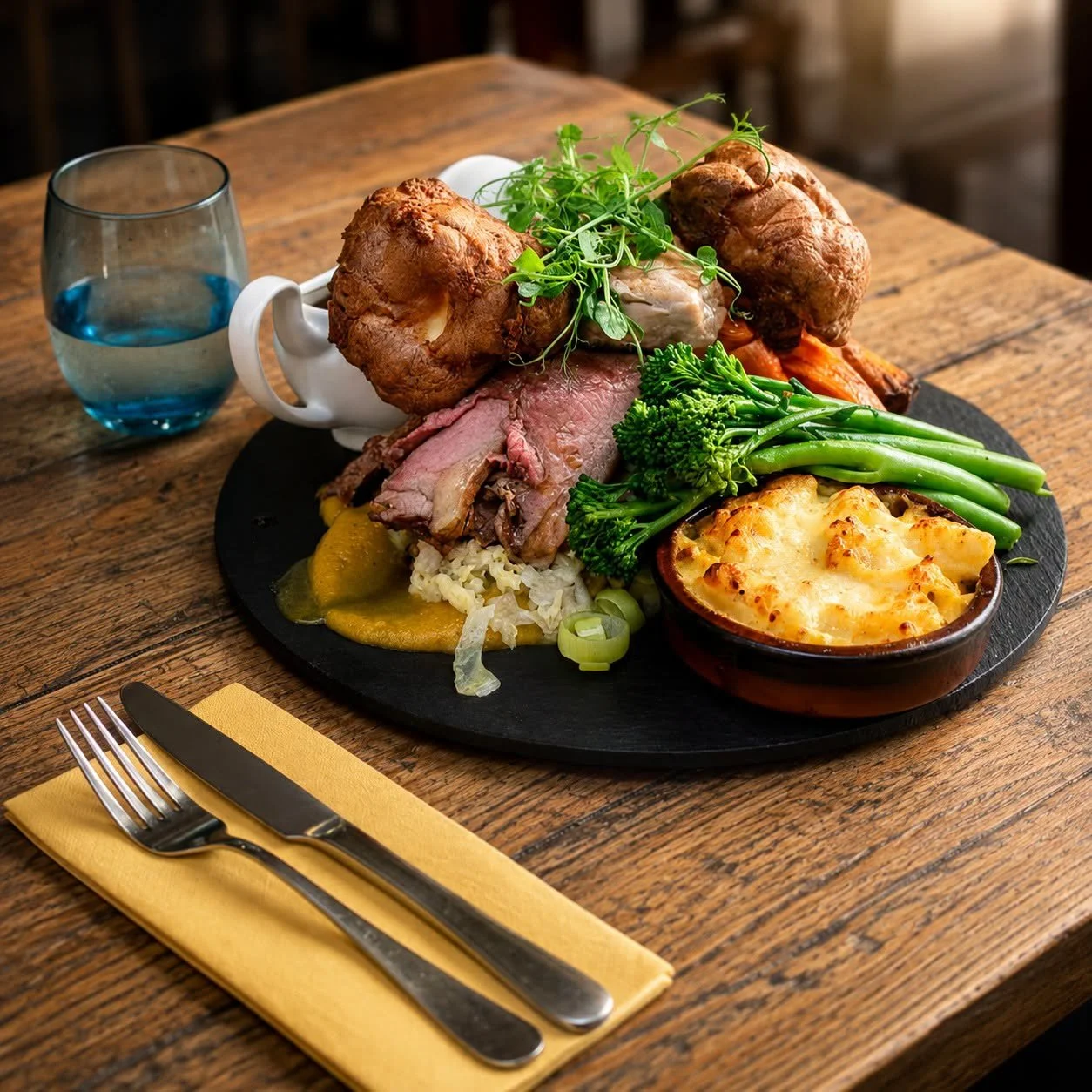 A plated meal with roasted chicken, sliced beef, green beans, broccoli, and a baked cheesy dish on a black round platter, accompanied by a glass of water, fork, and knife on a yellow napkin on a wooden table.
