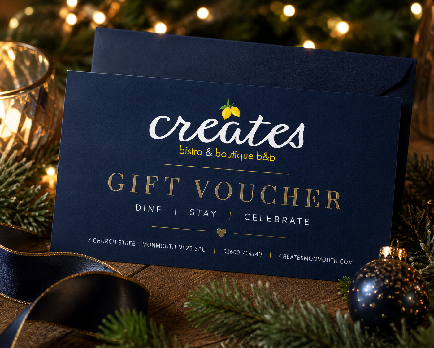 A navy blue gift voucher for 'creates bistro & boutique B&B' on a wooden surface, surrounded by Christmas greenery, a navy ribbon, and a blue Christmas ornament.