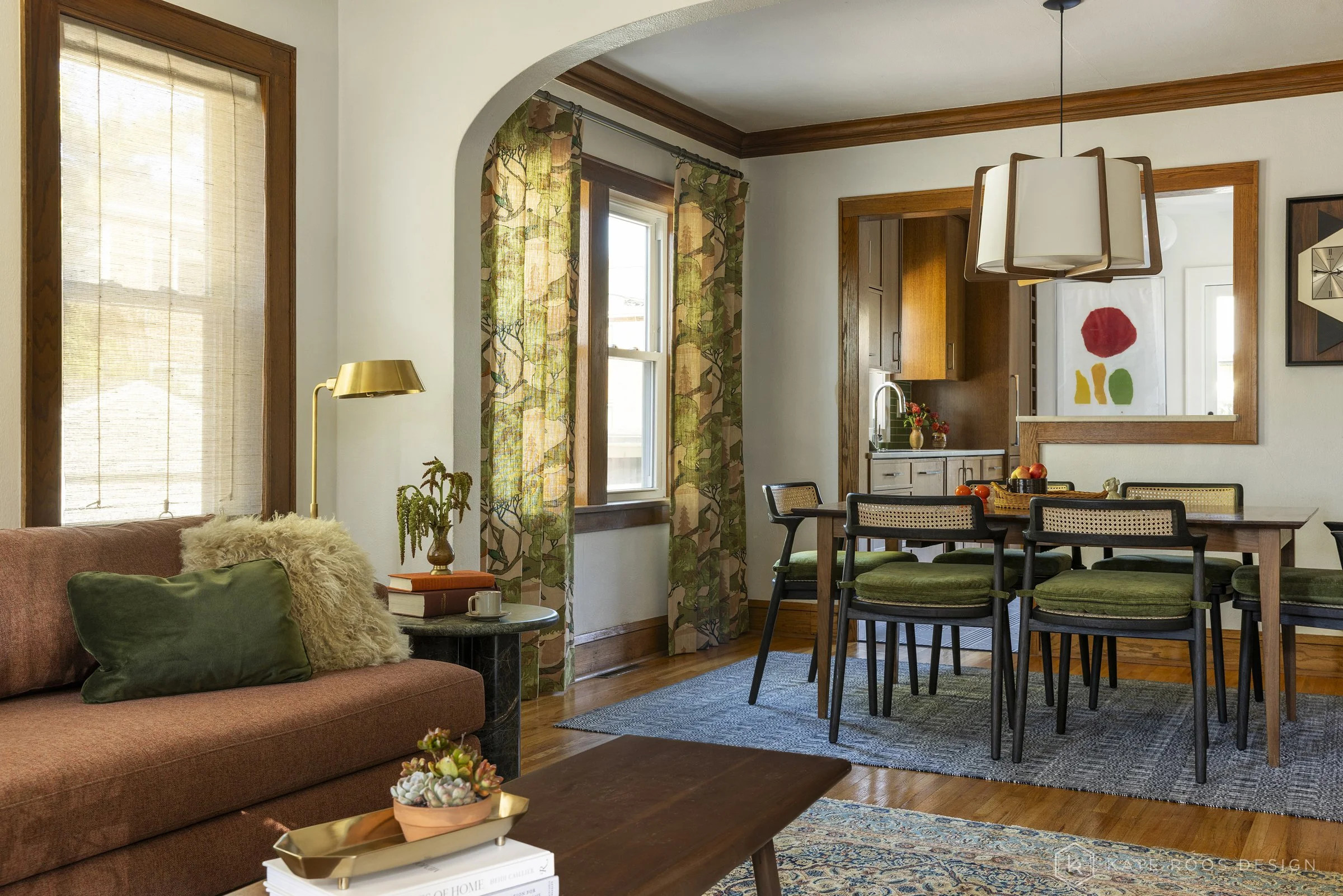 This curated living and dining room showcases a warm modern traditional interior design with strong mid-century modern influences. Original hardwood floors, rich stained wood trim, and a classic arched doorway highlight the home’s historic character 