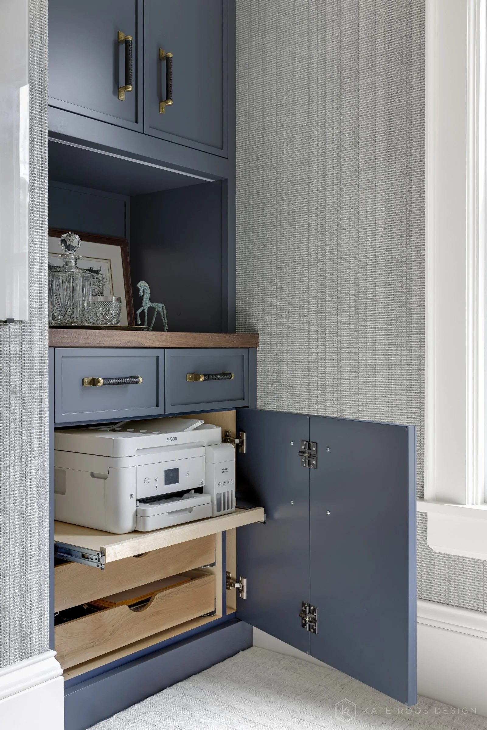 This image showcases a beautifully designed home office that balances function, comfort, and style. The space features a built-in desk with rich wood countertops and deep navy cabinetry accented by brass hardware, creating a sophisticated yet invitin