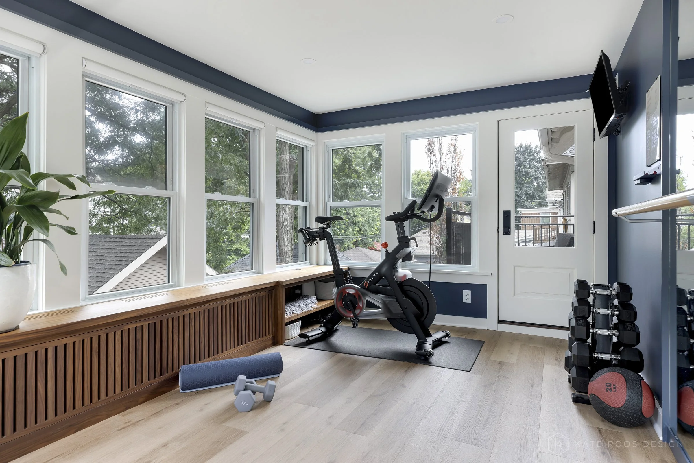 This image showcases a beautifully designed home office that balances function, comfort, and style. The space features a built-in desk with rich wood countertops and deep navy cabinetry accented by brass hardware, creating a sophisticated yet invitin