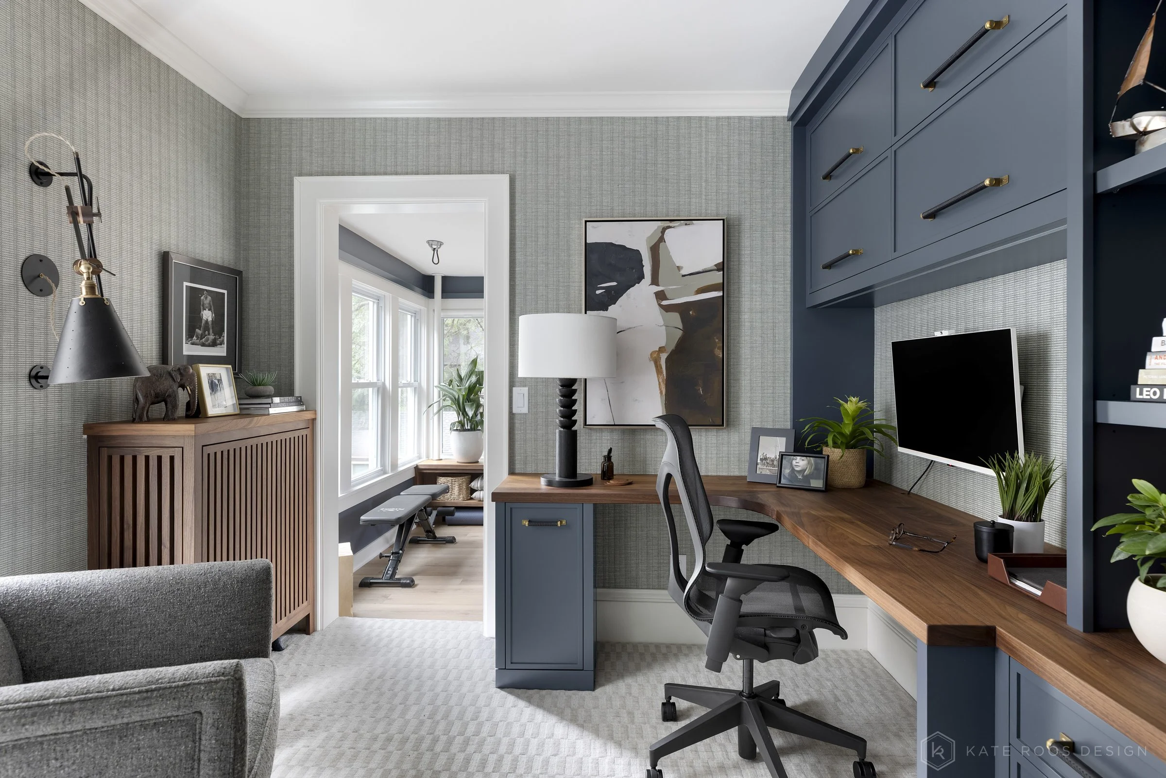 This image showcases a beautifully designed home office that balances function, comfort, and style. The space features a built-in desk with rich wood countertops and deep navy cabinetry accented by brass hardware, creating a sophisticated yet invitin
