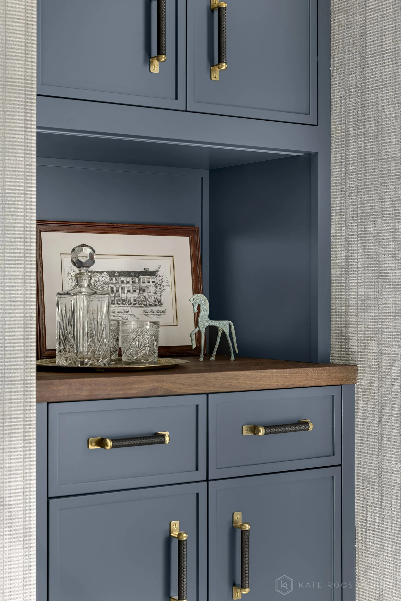This image showcases a beautifully designed home office that balances function, comfort, and style. The space features a built-in desk with rich wood countertops and deep navy cabinetry accented by brass hardware, creating a sophisticated yet invitin