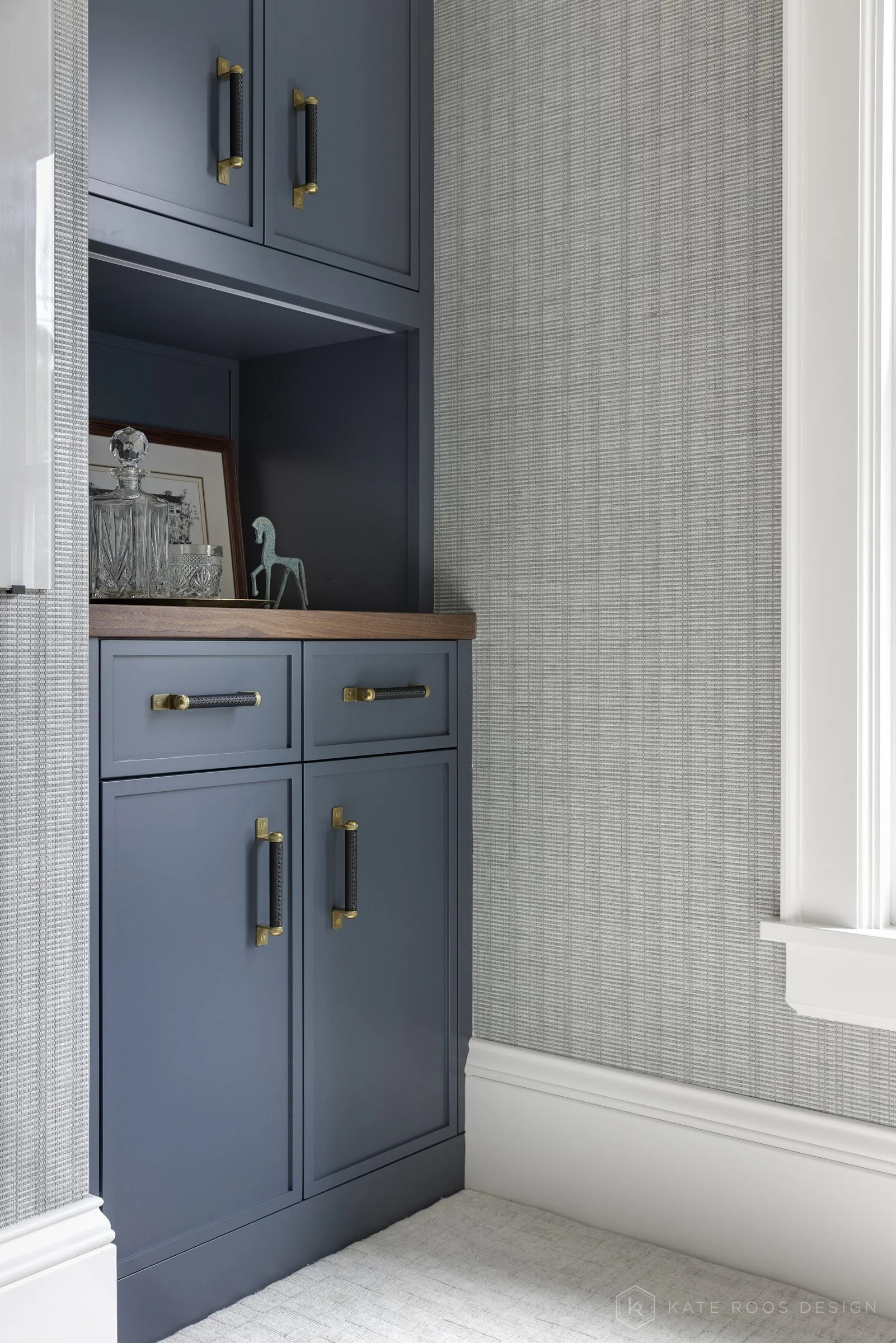 This image showcases a beautifully designed home office that balances function, comfort, and style. The space features a built-in desk with rich wood countertops and deep navy cabinetry accented by brass hardware, creating a sophisticated yet invitin