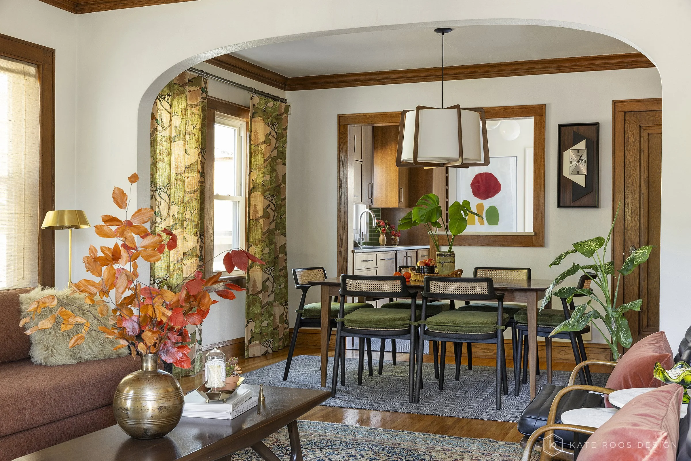 This curated living and dining room showcases a warm modern traditional interior design with strong mid-century modern influences. Original hardwood floors, rich stained wood trim, and a classic arched doorway highlight the home’s historic character 