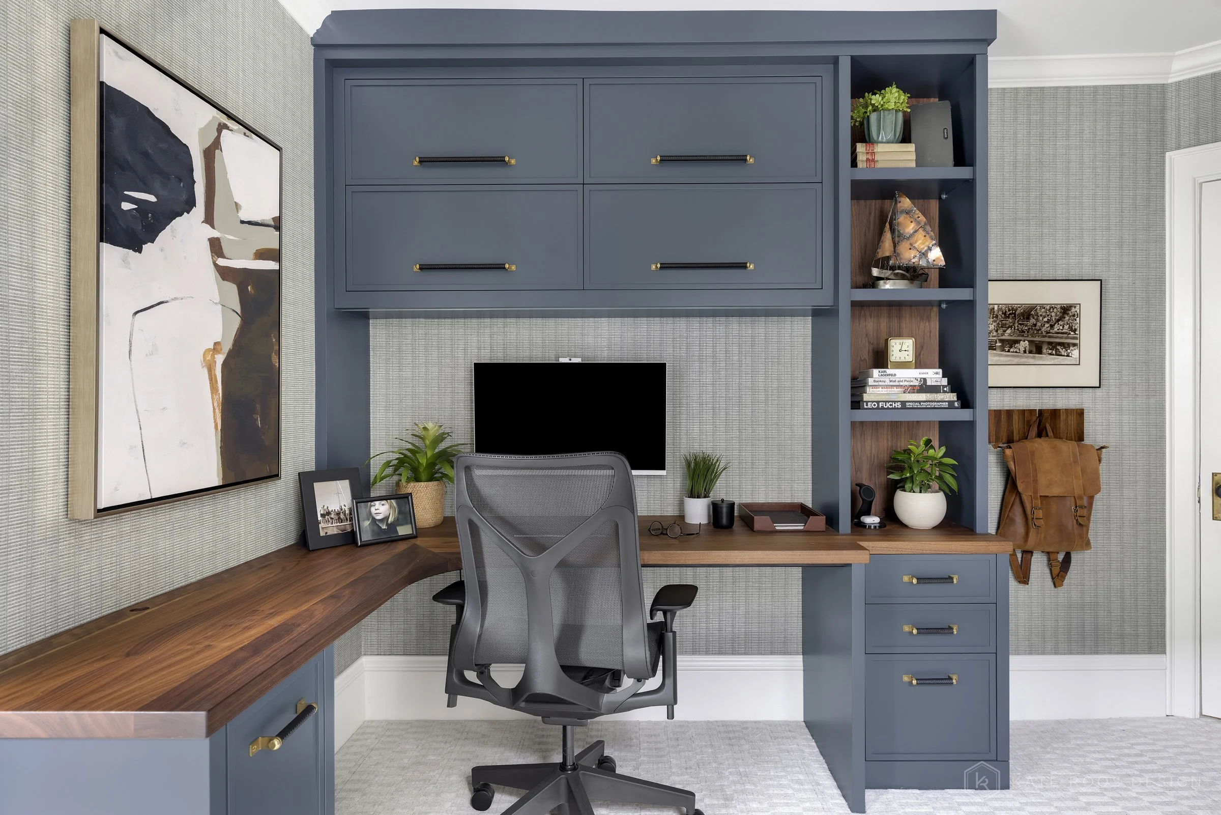 This image showcases a beautifully designed home office that balances function, comfort, and style. The space features a built-in desk with rich wood countertops and deep navy cabinetry accented by brass hardware, creating a sophisticated yet invitin