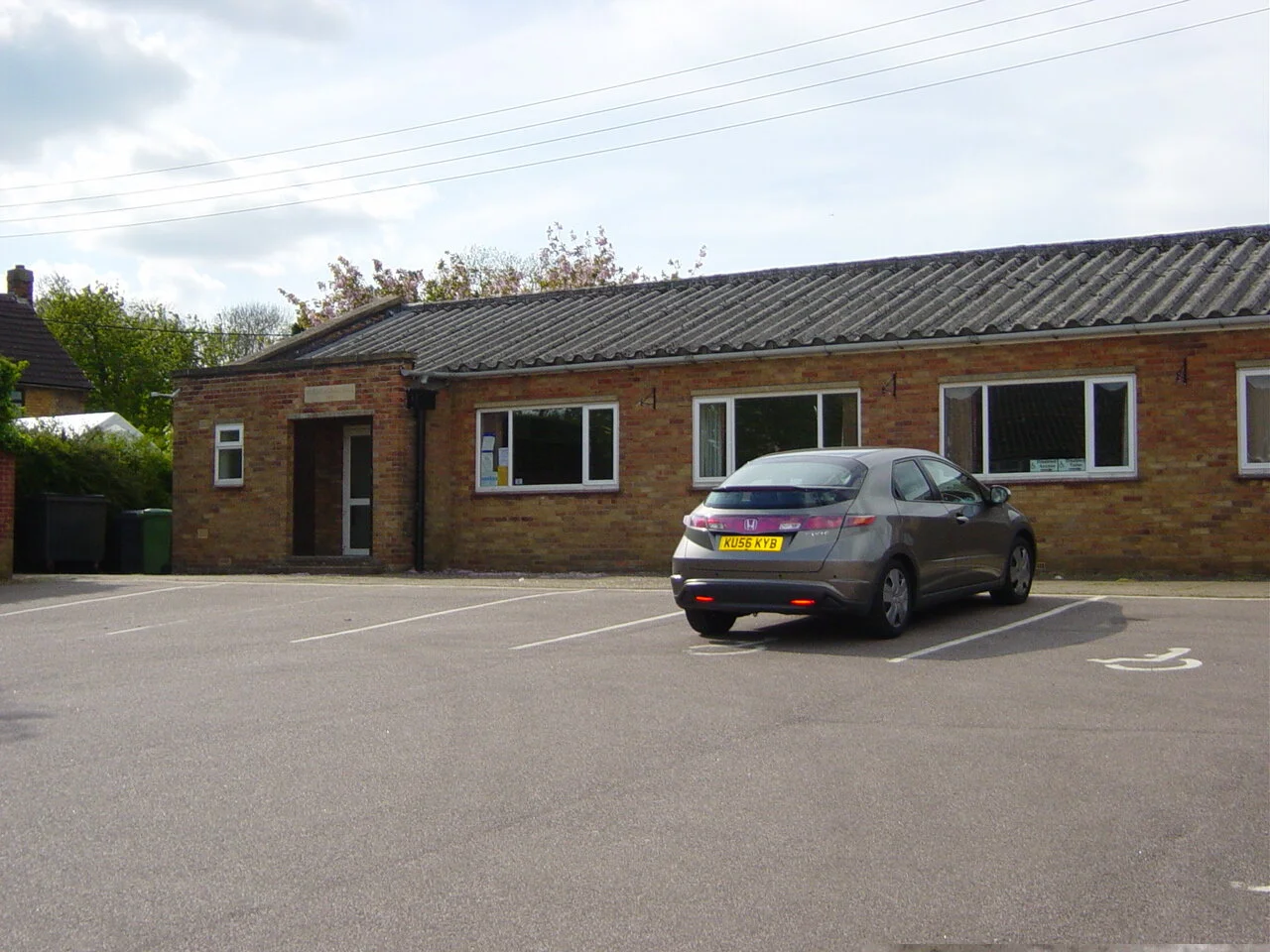 Brick building with a sloped roof and multiple windows, featuring a parked gray car in a mostly empty parking lot with a visible handicap parking space.