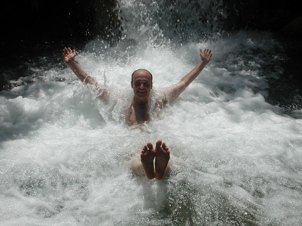Person sitting under a waterfall with arms raised, surrounded by splashing water, smiling.