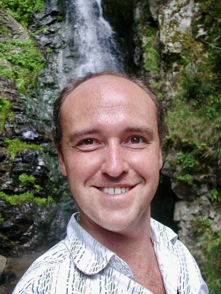 A person taking a selfie in front of a waterfall in a forested area.