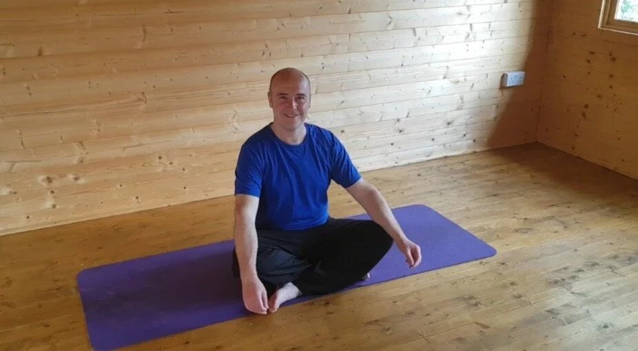Man in blue shirt sitting on purple yoga mat in wooden room