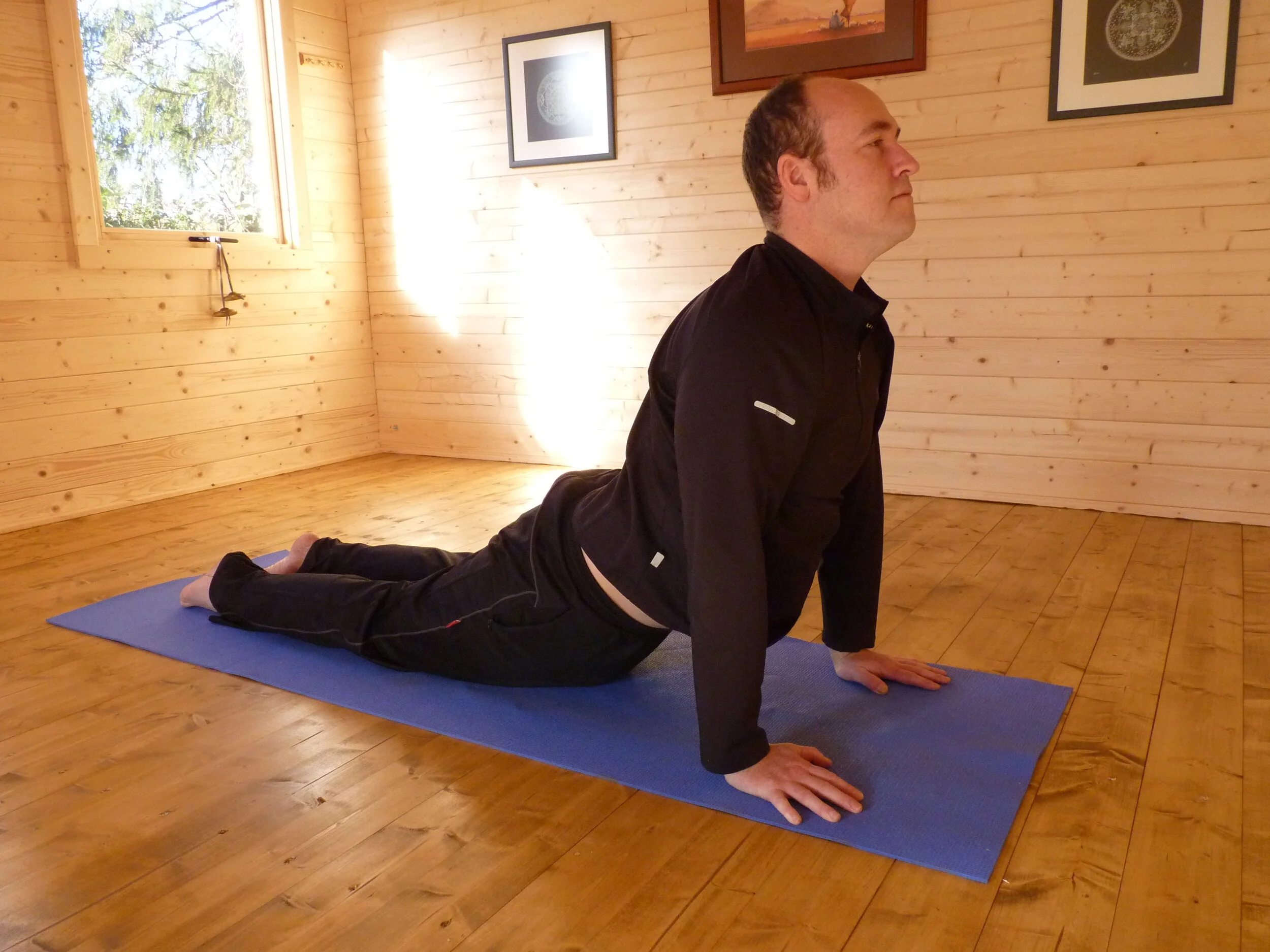 Person performing yoga pose on blue mat in wooden room with sunlight, framed pictures on wall.