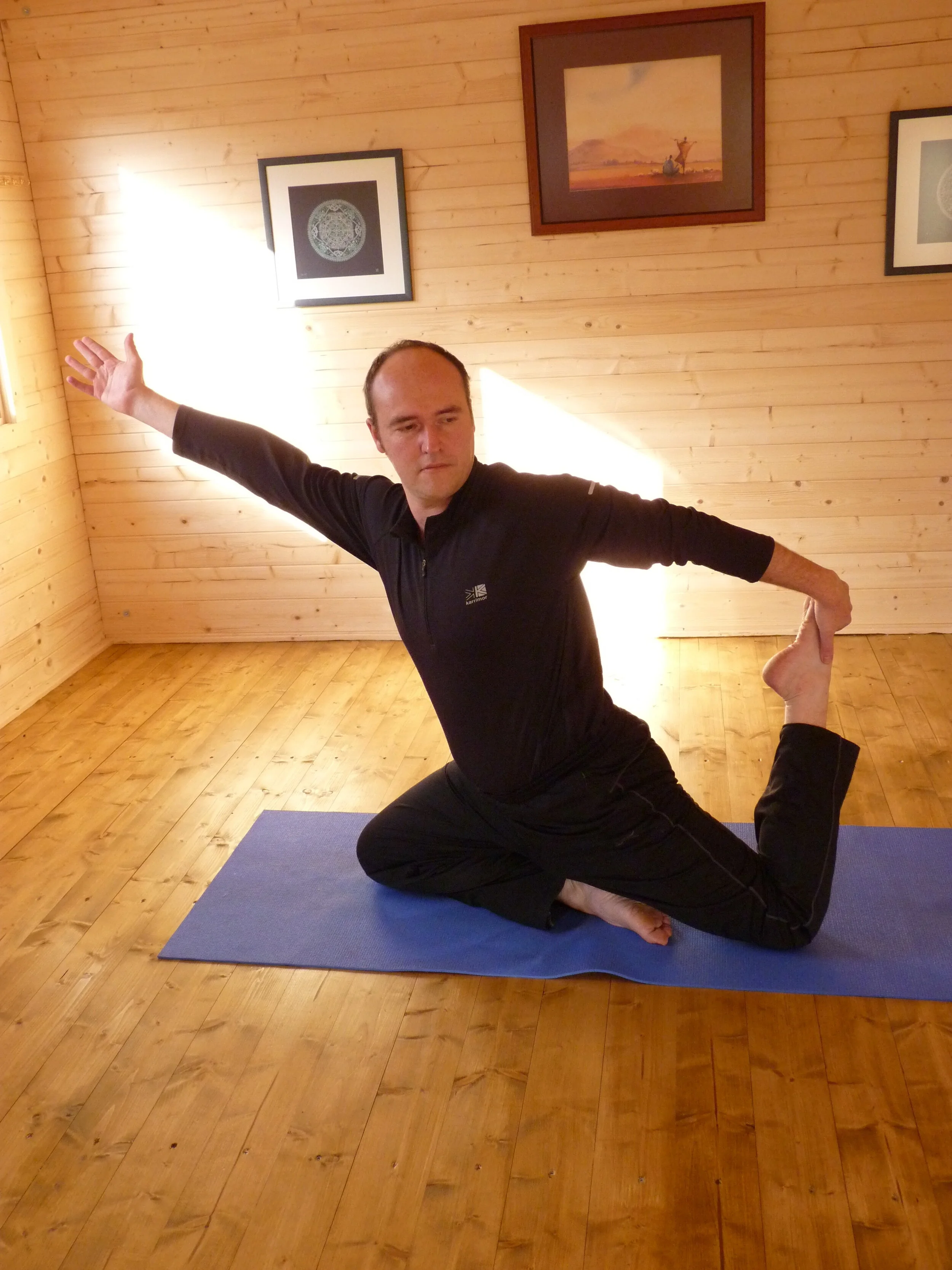 A person practicing yoga on a blue mat in a wooden room with framed artworks on the walls.