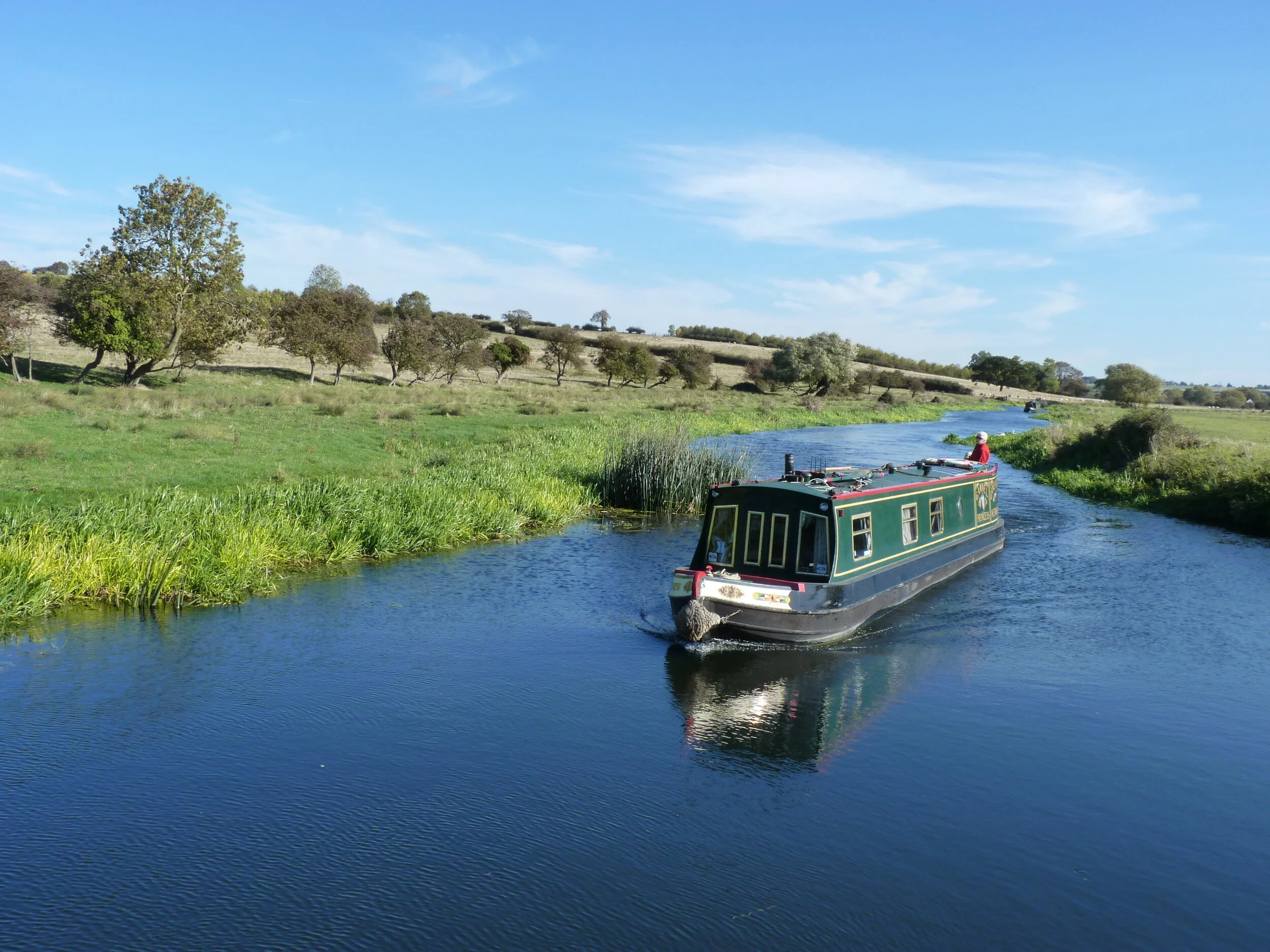 Narrowboat on a scenic river surrounded by green countryside and trees under a blue sky.