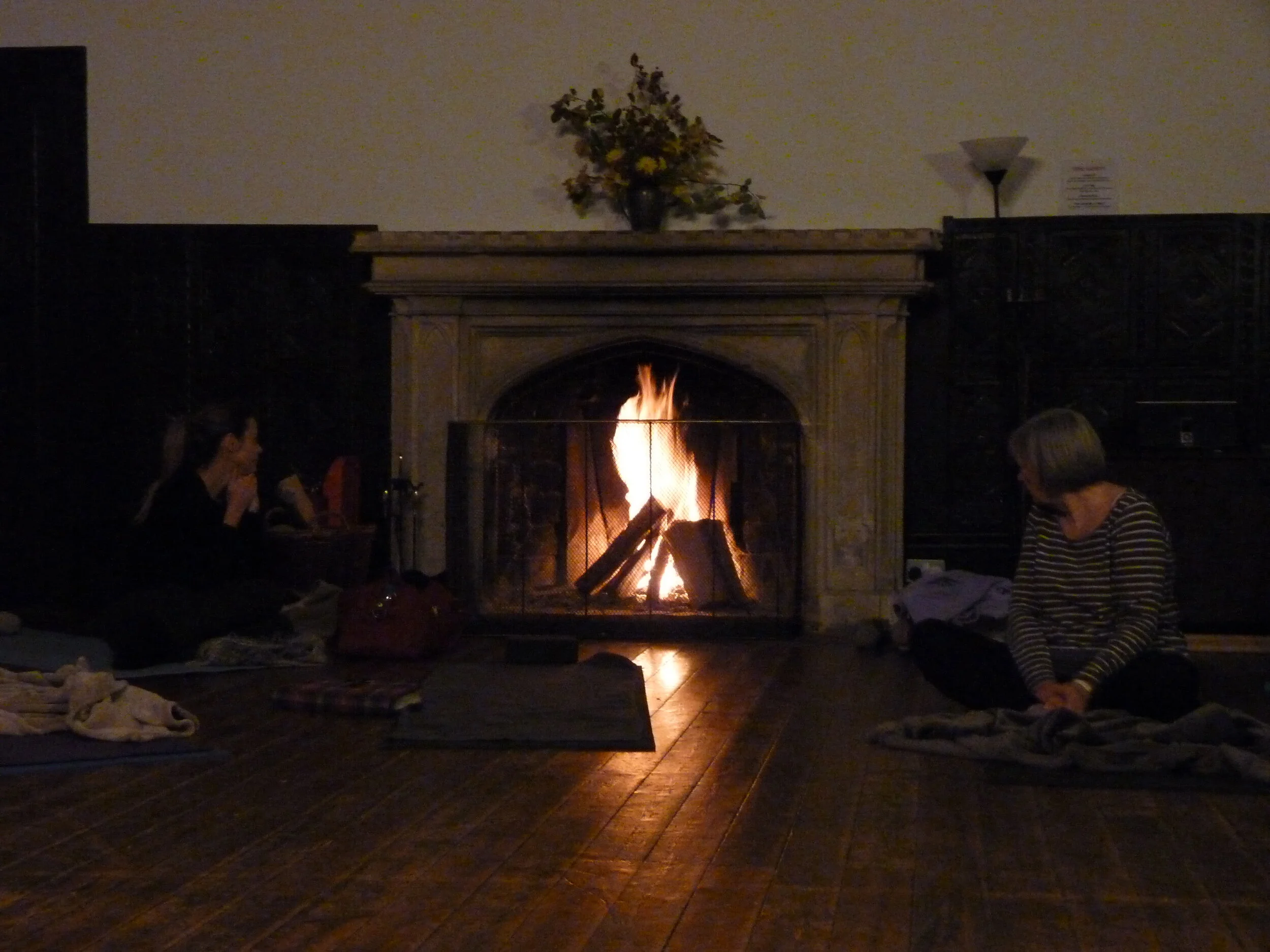 Two people sitting on the floor near a lit fireplace in a dimly lit room, with blankets and yoga mats.