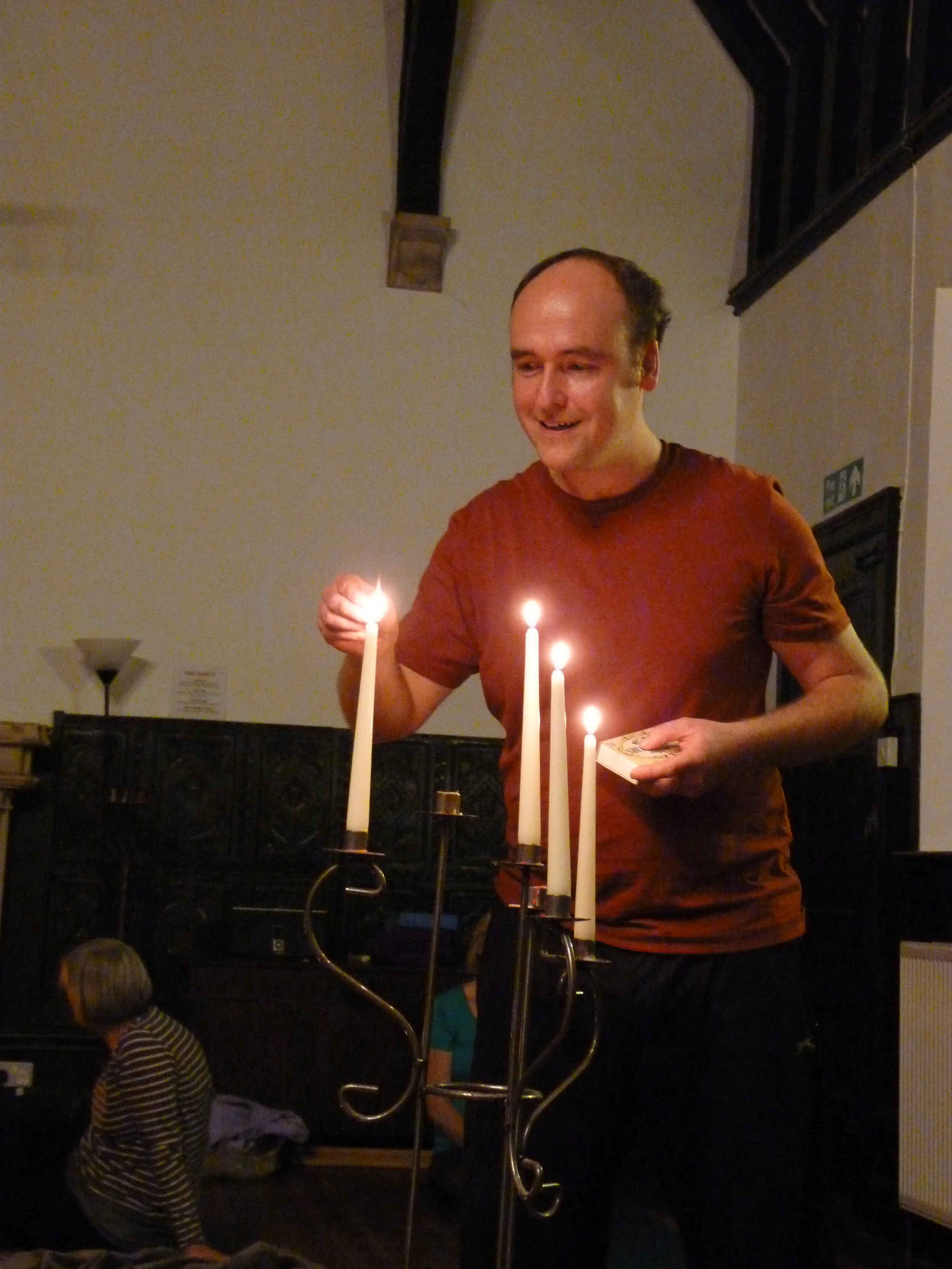 Man in red shirt lighting candles on a metal candelabra indoors.