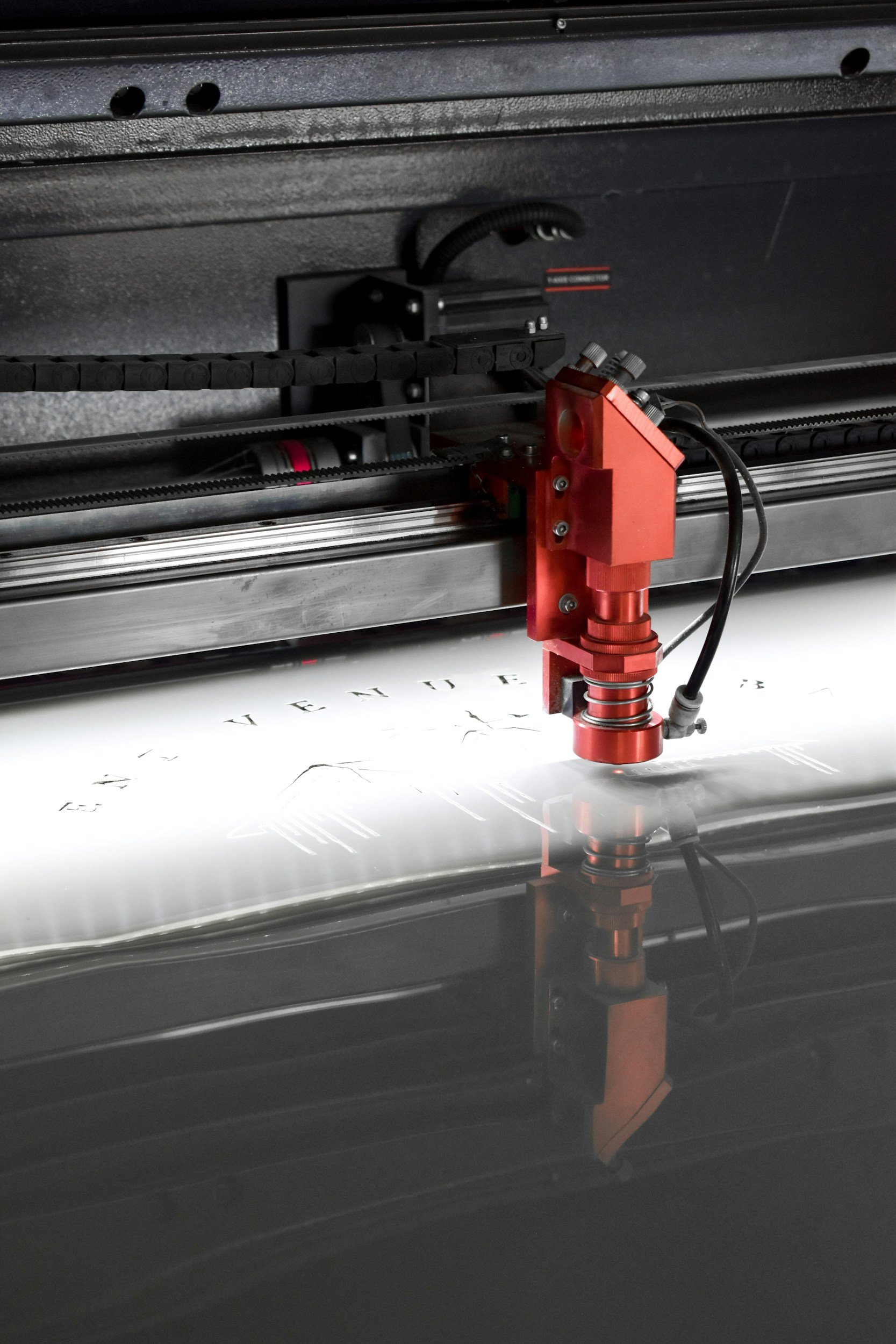 A close-up of a laser cutter engraving text and designs onto a reflective surface. The laser head is red, positioned near the center, with a black cable attached. The surface reflects part of the laser and the engraved designs.