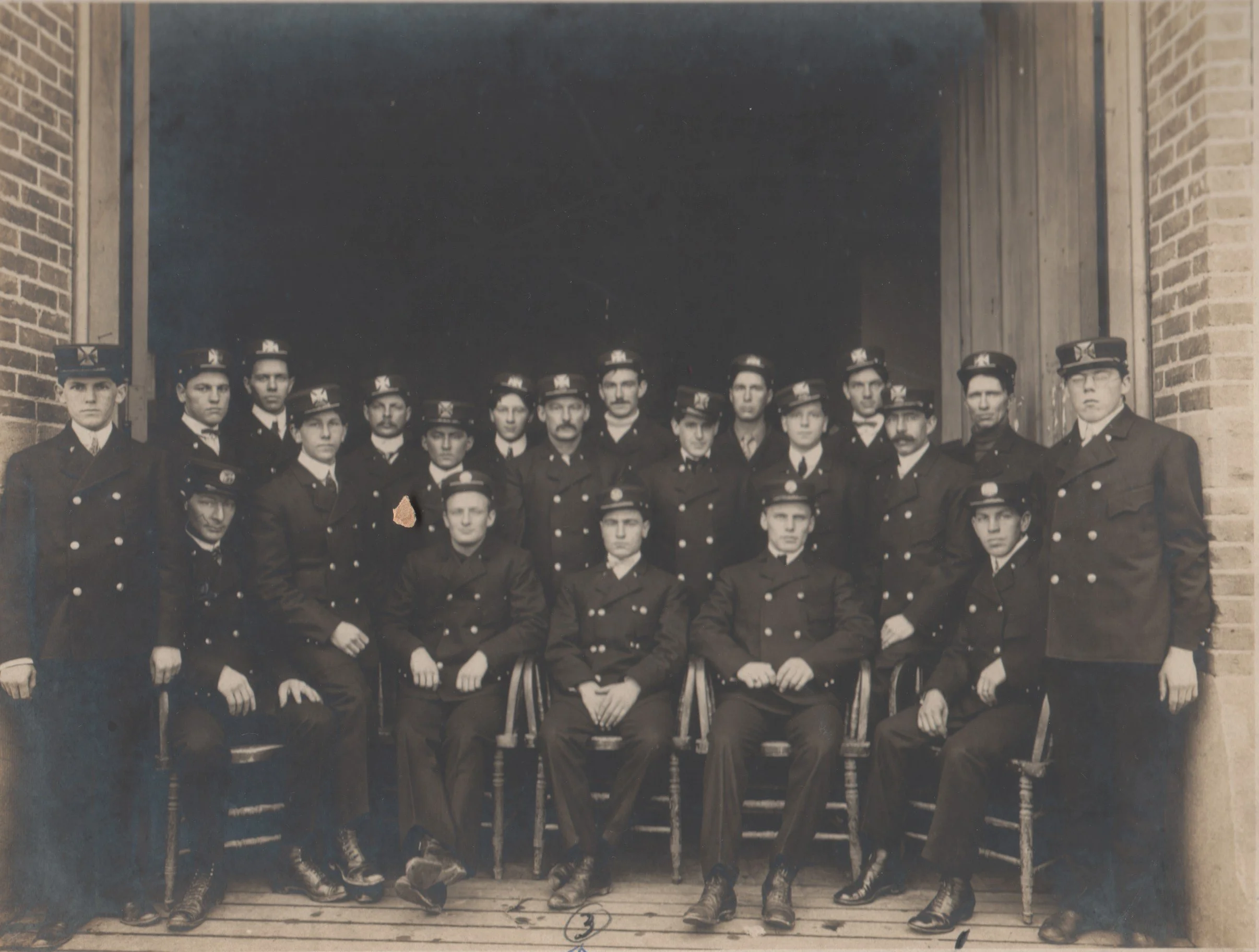  Group photograph of 21 members of the 1907 Anacortes Fire Department in uniform. Picture is taken in the doorway of the firehouse. Seated left to right: 1) George Rodenbaugh, 3) J. Elmer Amsberry, Fire Chief, 5) Aubrey Sumner. Man standing at extrem