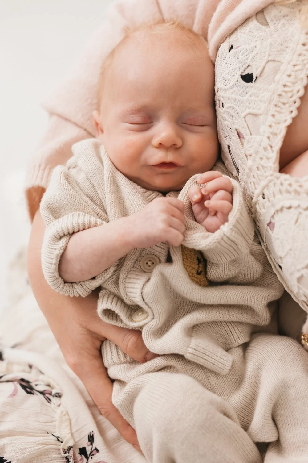 A sleeping baby held in a woman's arms, wearing a beige sweater and pants, with hands clasped together, close-up of peaceful face and closed eyes.