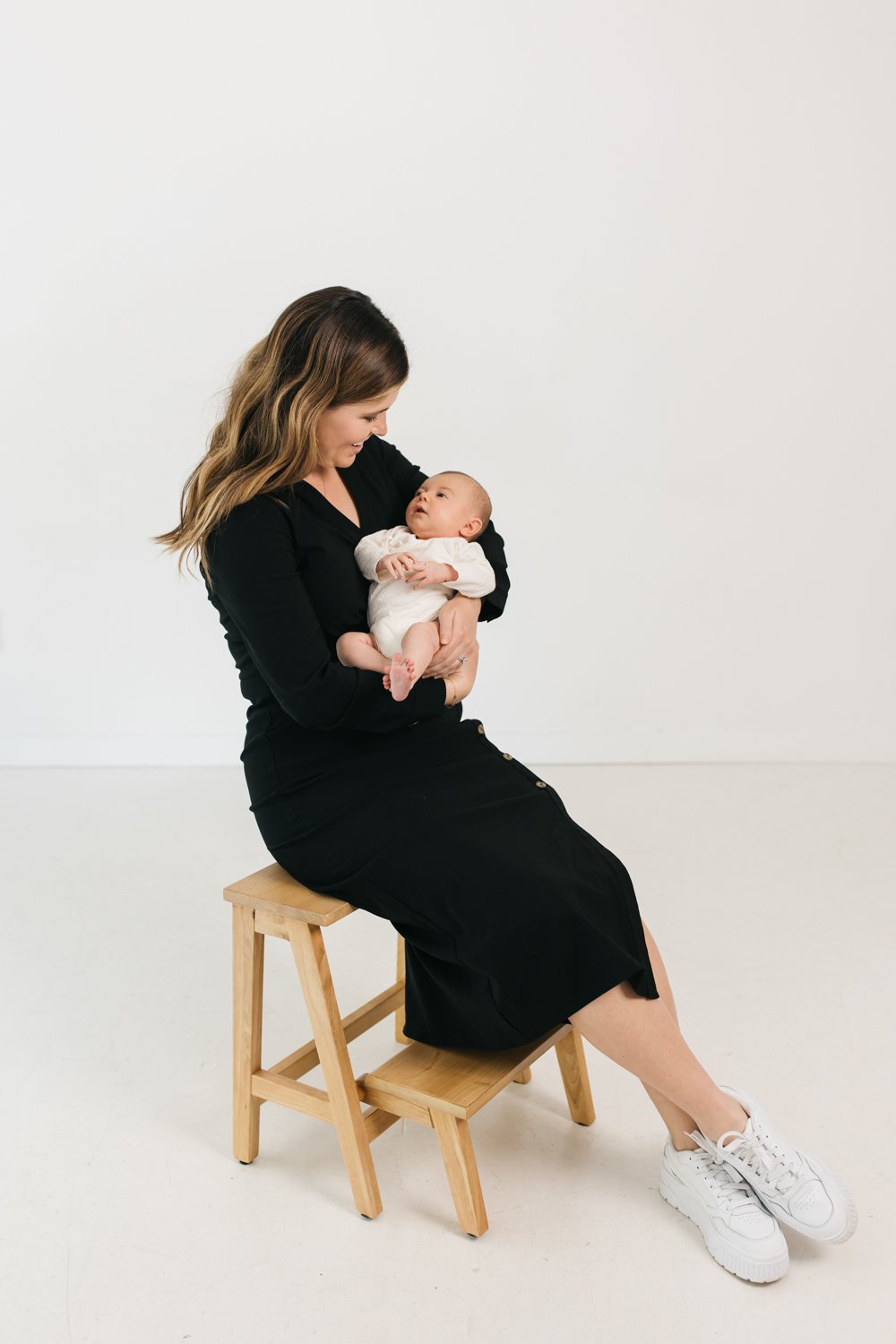 A woman in a black dress and white sneakers sitting on a small wooden chair, holding a baby in her arms, looking at the baby with a gentle smile against a plain white background.