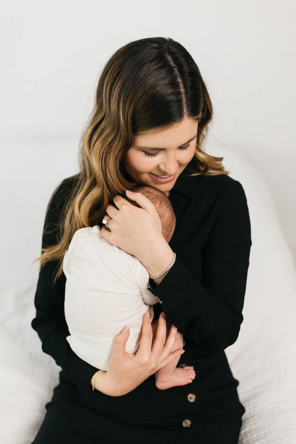 A woman with long, wavy brown hair holding a newborn baby close, smiling and looking down affectionately, against a plain white background.