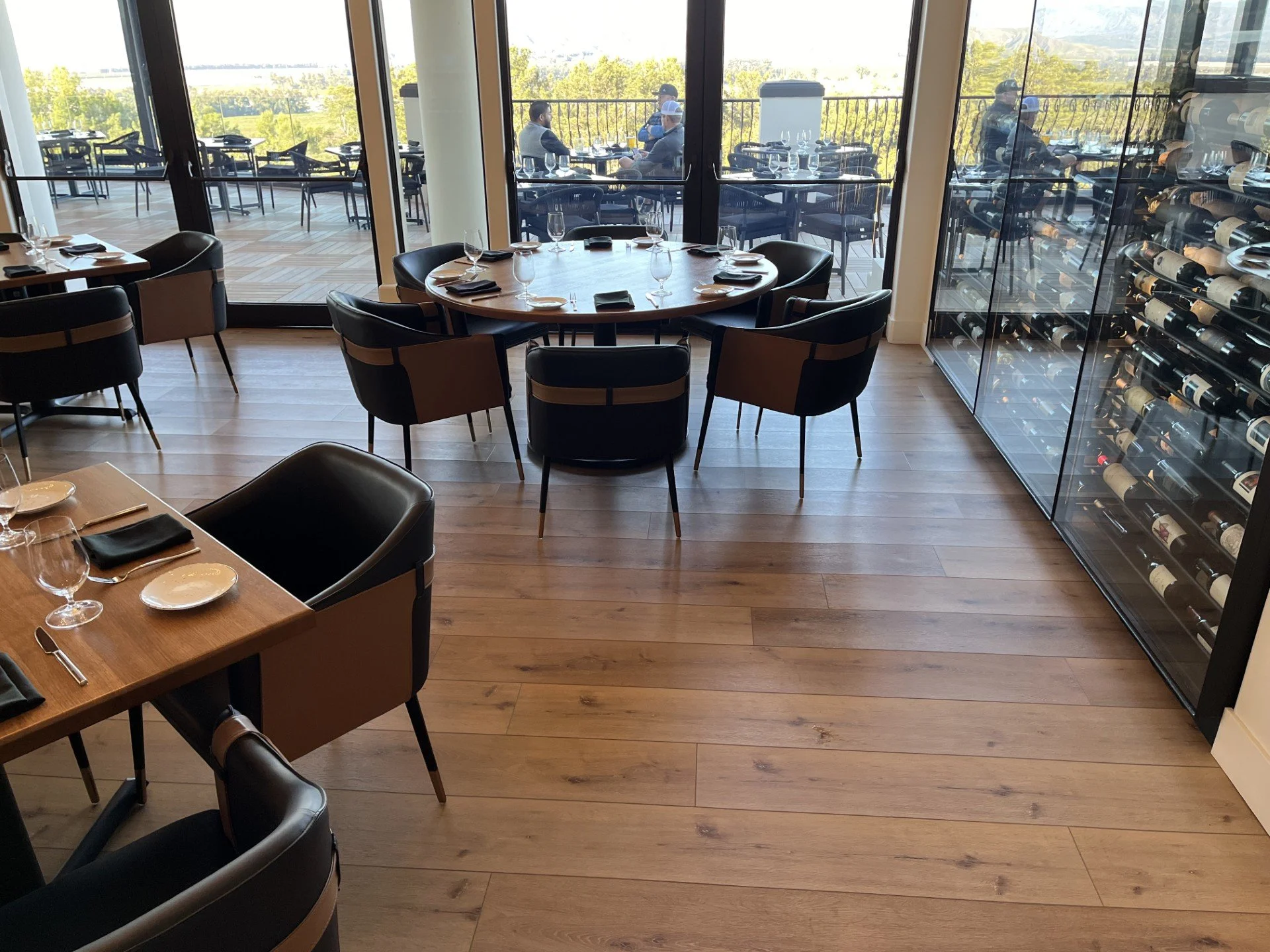 Interior of a restaurant with a round table set for dining, black chairs surrounding it, and a glass wine fridge on the right. Large glass doors lead to an outdoor balcony with additional seating and a scenic view of trees and a body of water in the 