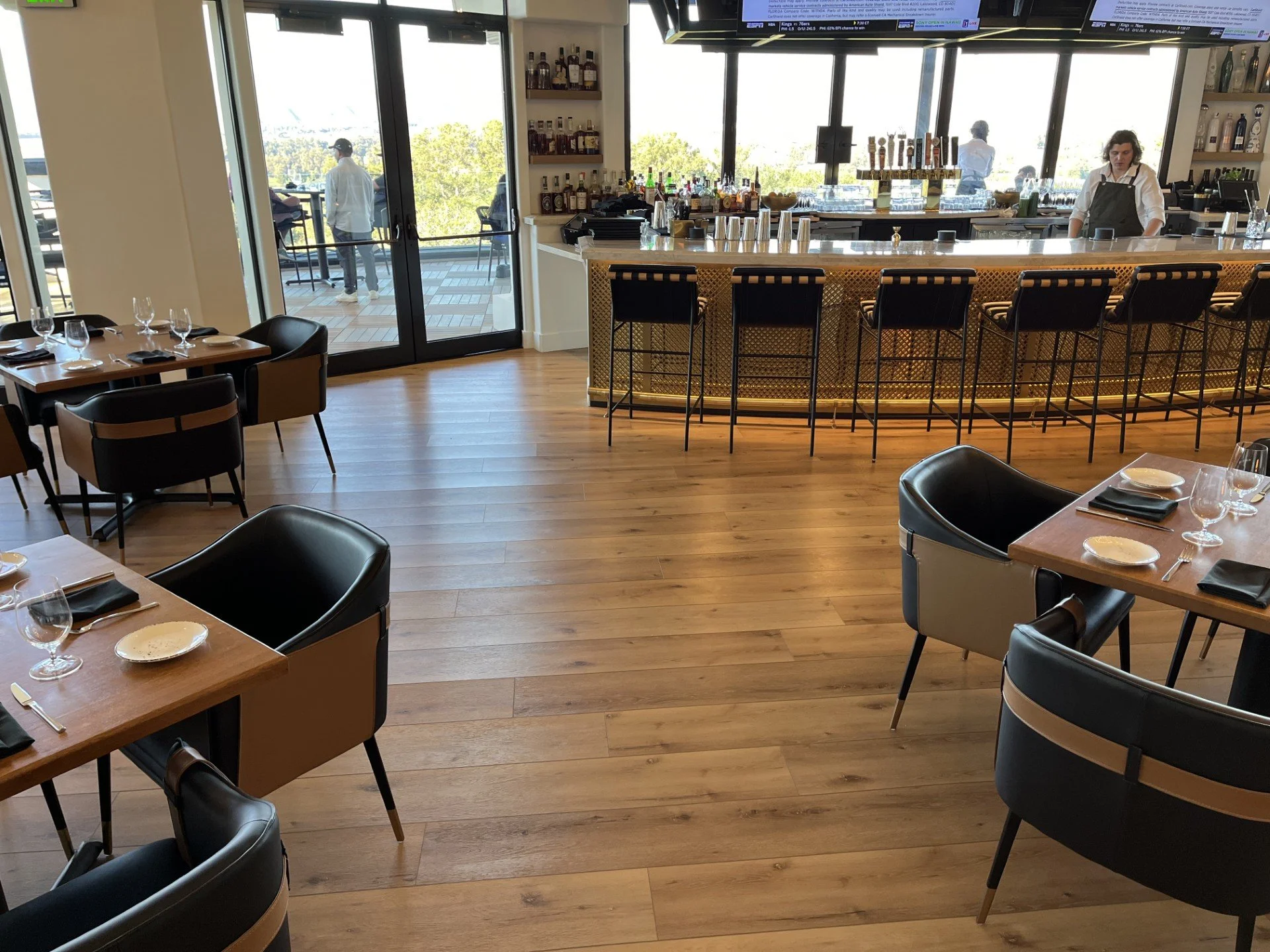 Interior of a restaurant with tables, black chairs, and a bar area with barstools. Glass doors lead outside where a person is walking on a balcony. The restaurant is well-lit with large windows and a TV screen is visible at the top of the image.