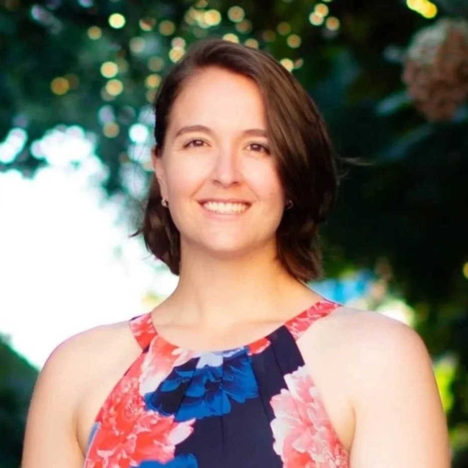 Smiling dietitian with brown hair standing outdoors, wearing a light-colored jacket and white top, with green foliage in the background.