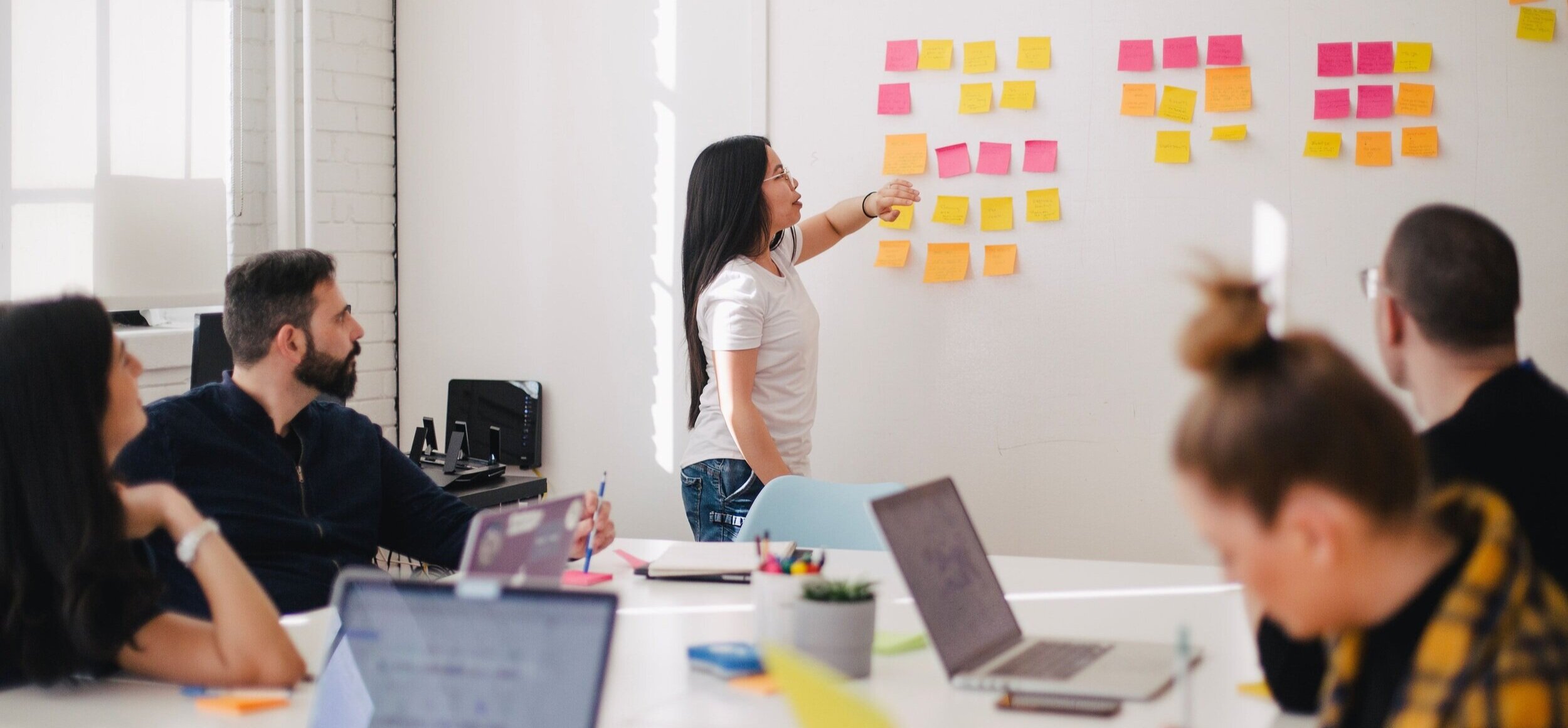 A woman presenting in front of a whiteboard with colorful sticky notes, while five colleagues seated at a table watch and take notes in a bright office.