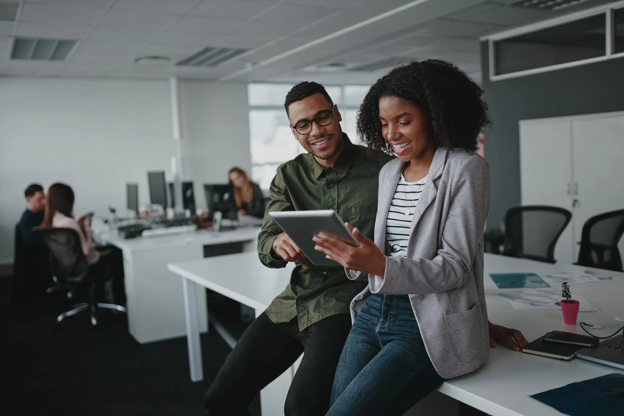 Two colleagues smiling and looking at a tablet in a modern office, with other employees working at desks in the background.