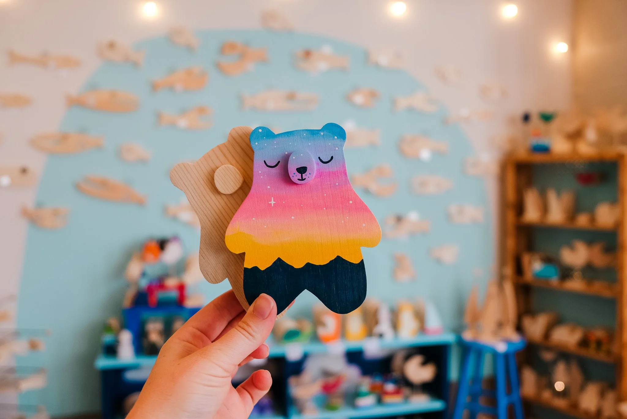 A hand holding a wooden bear-shaped toy with a colorful rainbow gradient face and closed eyes in a room decorated with a fish mural and nautical-themed decorations.