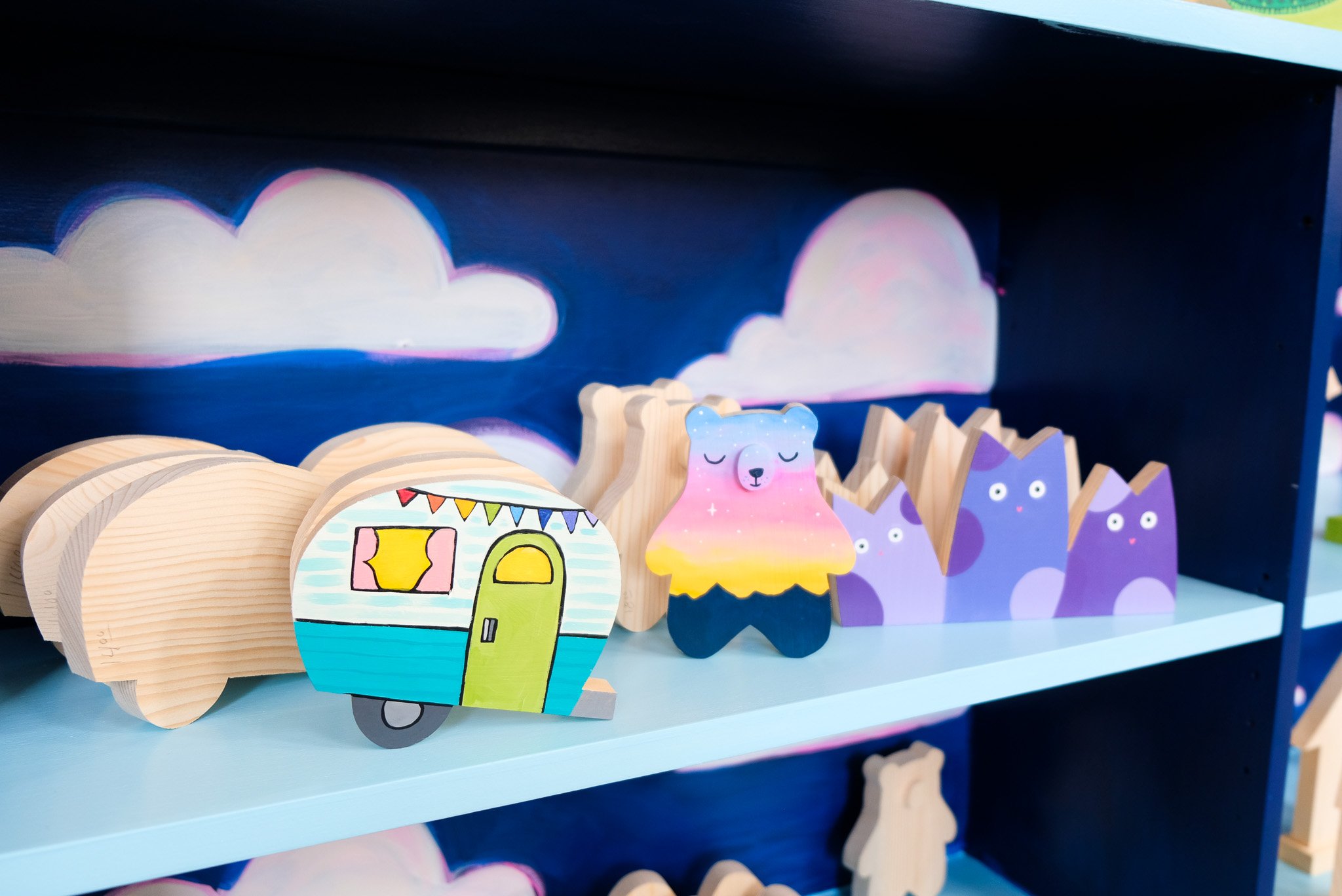 Colorful wooden toys on a blue shelf with painted clouds and pink ray sky background.