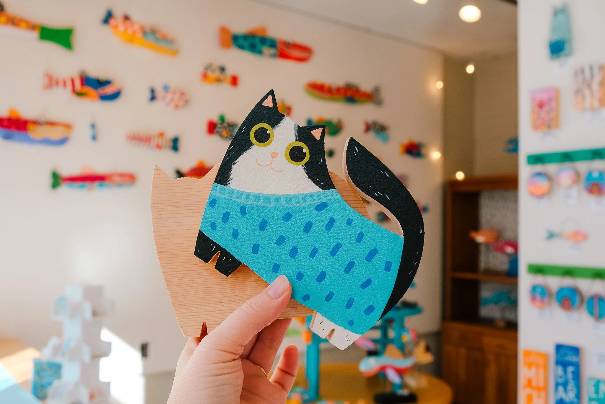A person holding a colorful paper craft of a black and white cat wearing a blue sweater, with a background of various paper fish decorations on the wall.