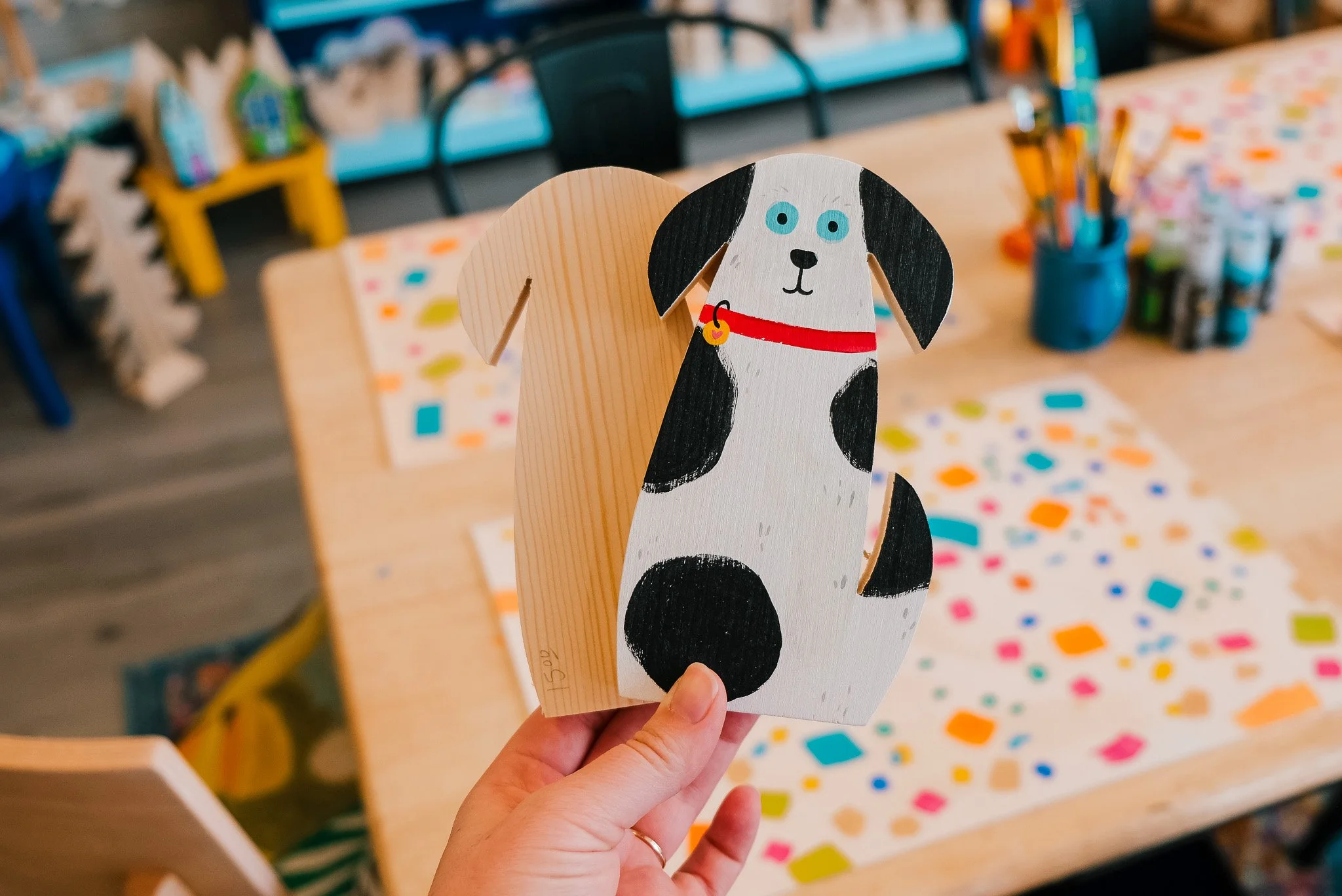 A hand holding a painted wooden dog figure with black and white spots, blue eyes, and a red collar in a colorful art classroom.