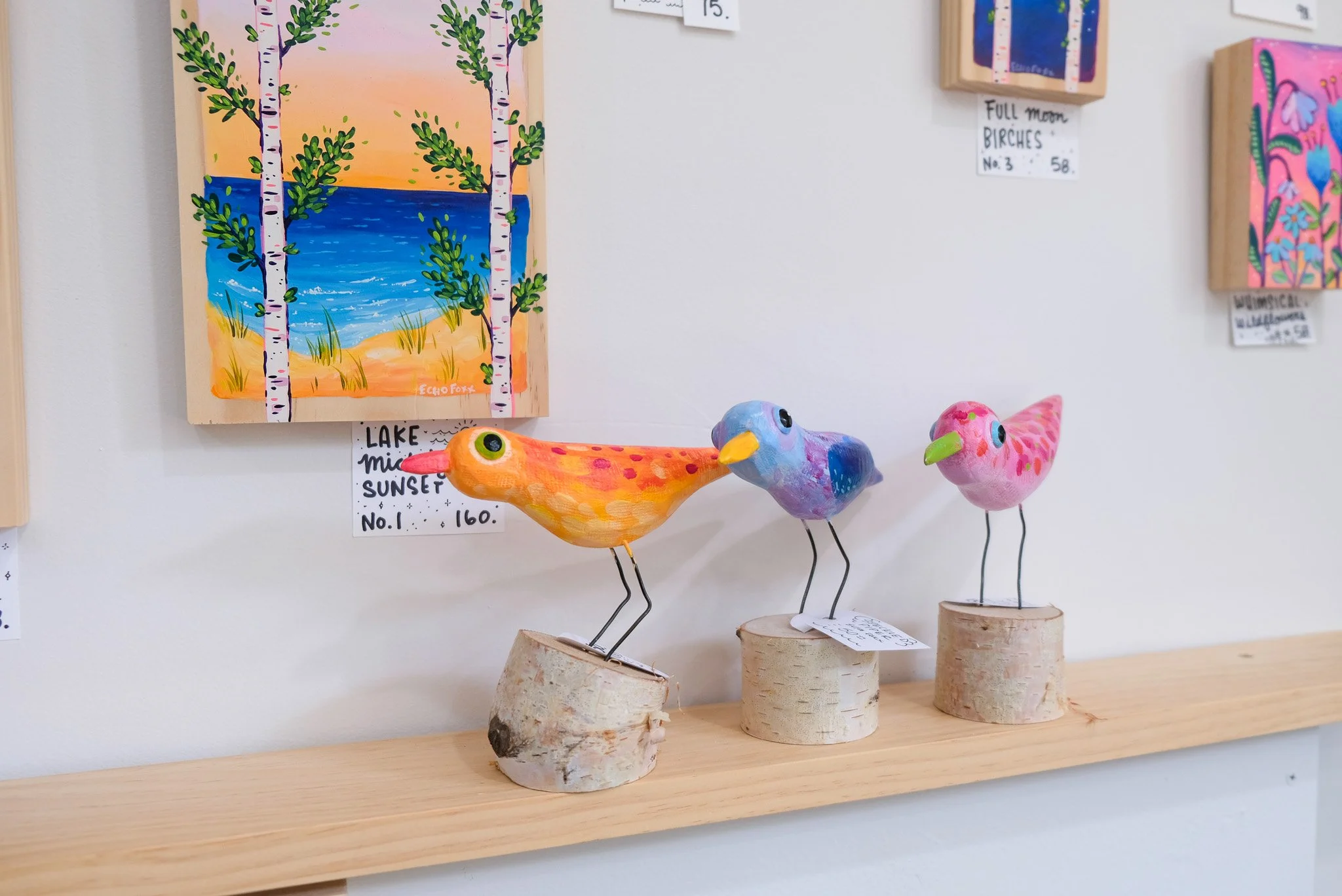 Decorative arts and crafts display including three colorful bird sculptures on wooden bases, with hand-painted landscape and artwork on the wall behind.