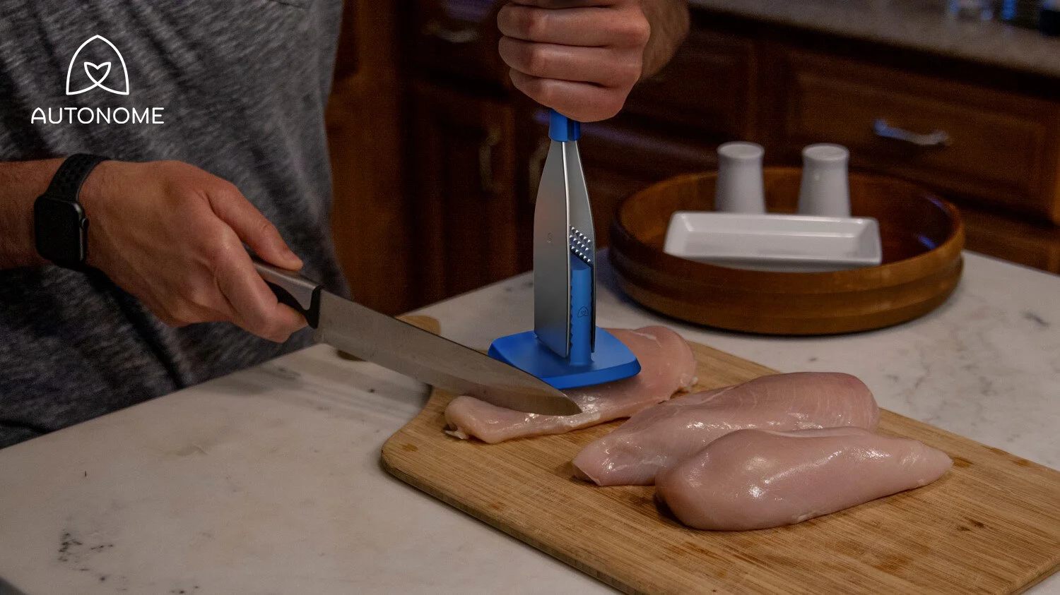A person slicing raw chicken breasts on a wooden cutting board with a knife. There is a manual meat slicer holding a chicken breast. In the background, there are white mugs in a wooden tray.