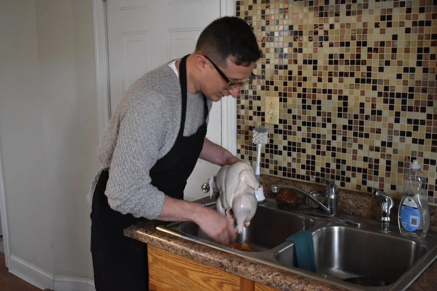 A man wearing glasses, a gray sweater, and a black apron is cleaning a raw turkey in a kitchen sink.