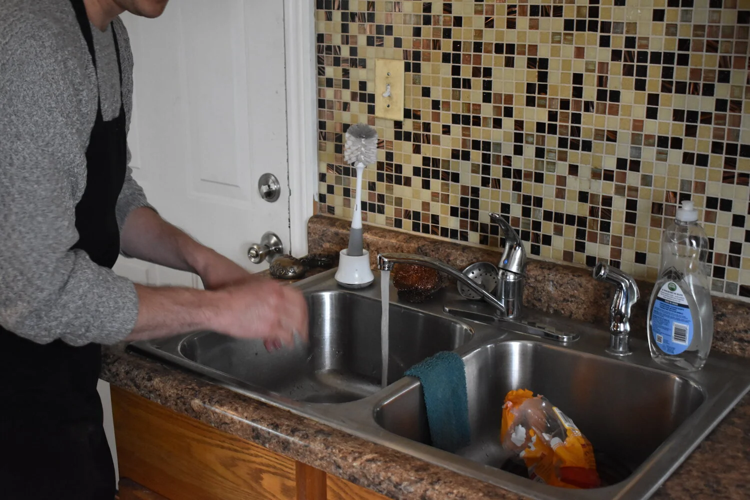 Person washing dishes at a kitchen sink with soap and a dish sponge, with a mosaic tile backsplash in the background.