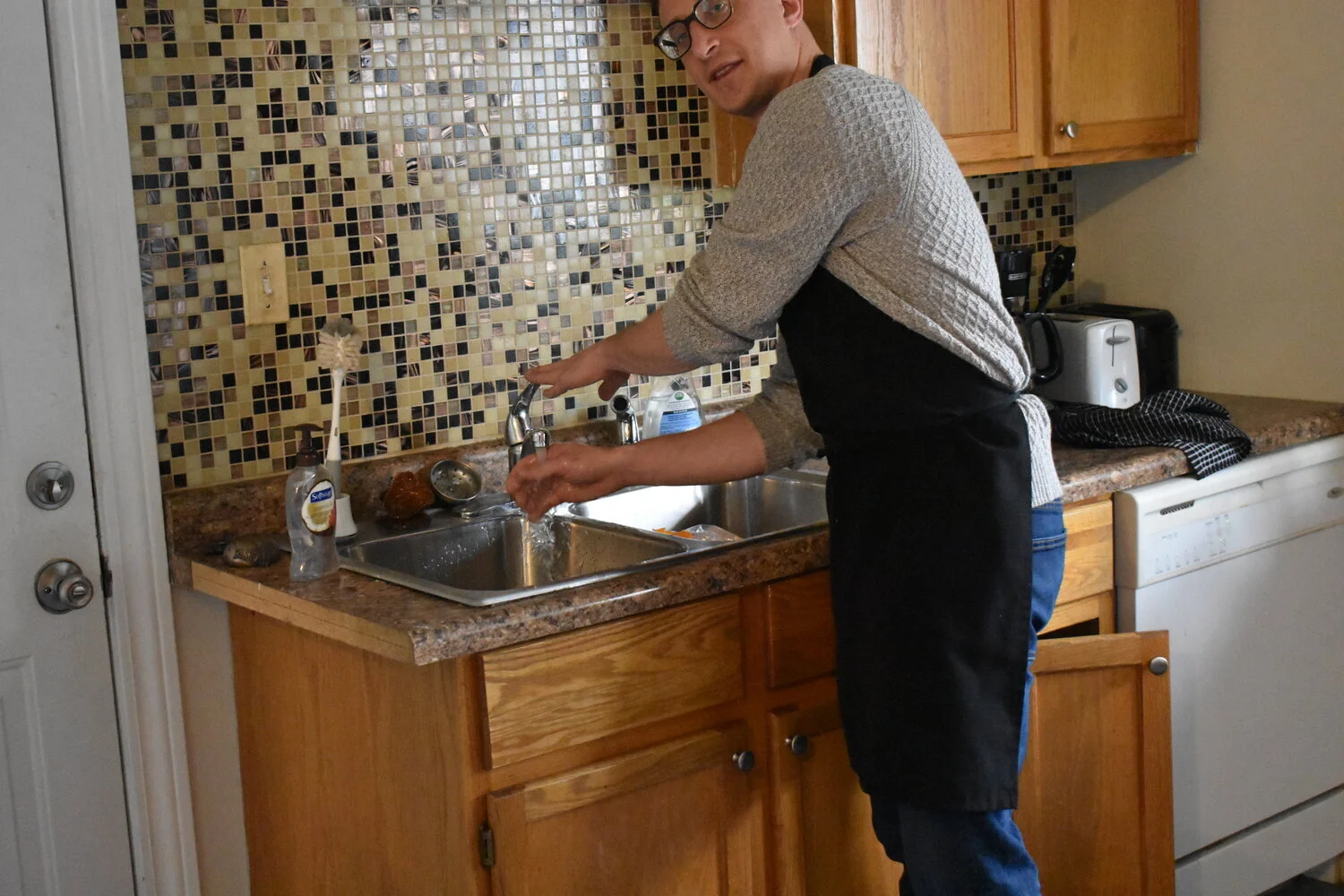 A man in glasses and a gray sweater washing dishes at a kitchen sink.