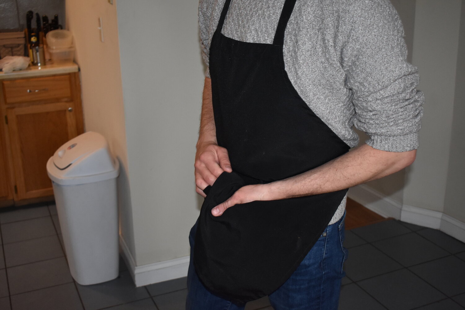 Person wearing a gray sweater and black apron in a kitchen, with a trash can visible in the background.