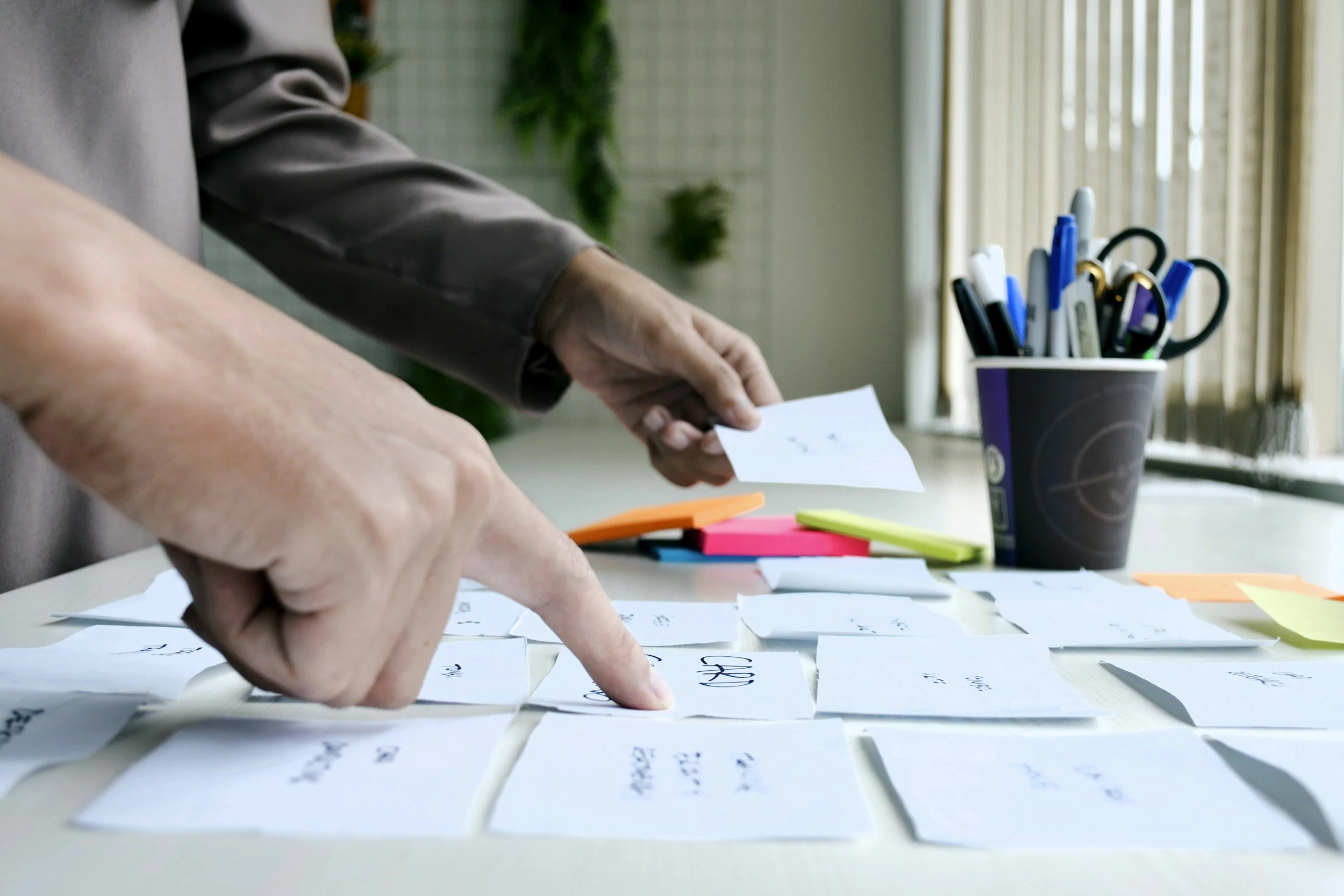 Person organizing papers on a desk, with a cup holding pens and markers nearby, in a well-lit room.