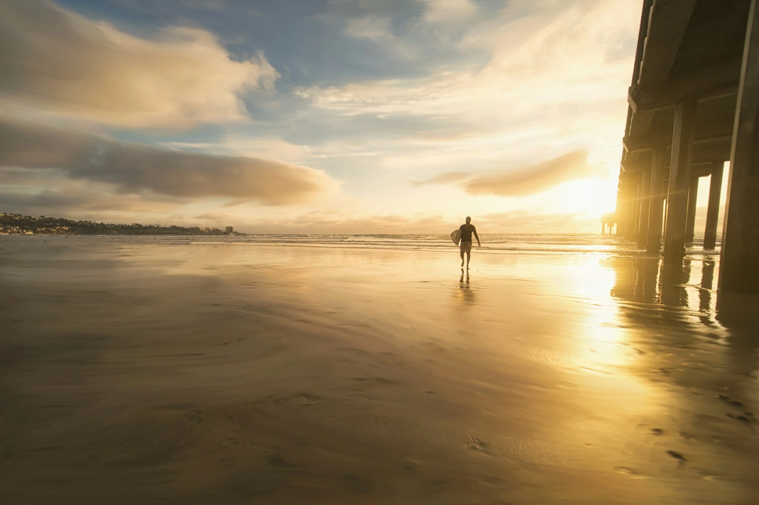 A person walking along the wet shoreline under a pier at sunset, with the sky partly cloudy and the sun setting on the right.